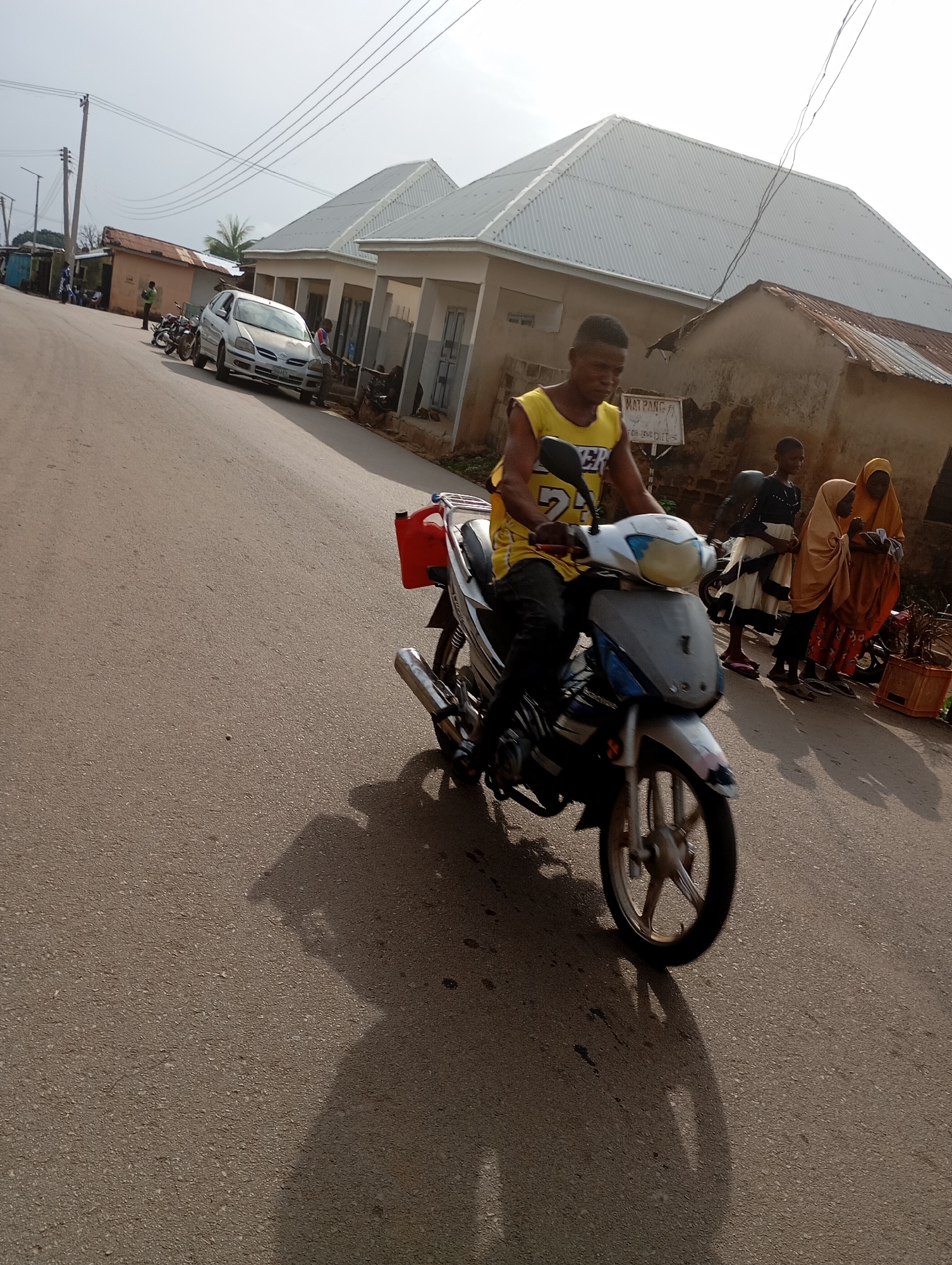 A young man is riding a white and silver motorcycle with a red jerrycan strapped to the rear rack on a paved road in Shendam, Nigeria. He is wearing a yellow tank top with the number "24" on it and black pants. Behind him, three females are standing on the side of the road. The woman closest to the motorcycle wears a dark top and a white skirt. The second woman wears a light brown hijab and a patterned skirt. The third woman, on the far right, wears a dark orange hijab and a patterned skirt. To the left of the road, a silver car is parked. Several buildings line the street, some with corrugated metal roofs and others with lighter-colored, sloping roofs. The sky is bright and appears overcast. A utility pole with overhead wires is visible on the left side of the image.