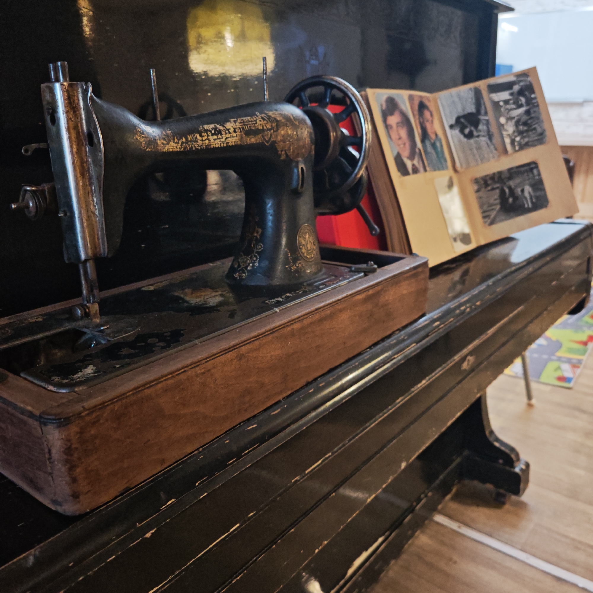 An antique sewing machine sits on a black piano in a room in Rostov, Russia. A partially open photo album rests on the piano next to the sewing machine. A child's play mat is visible in the background on a light-colored wood floor.