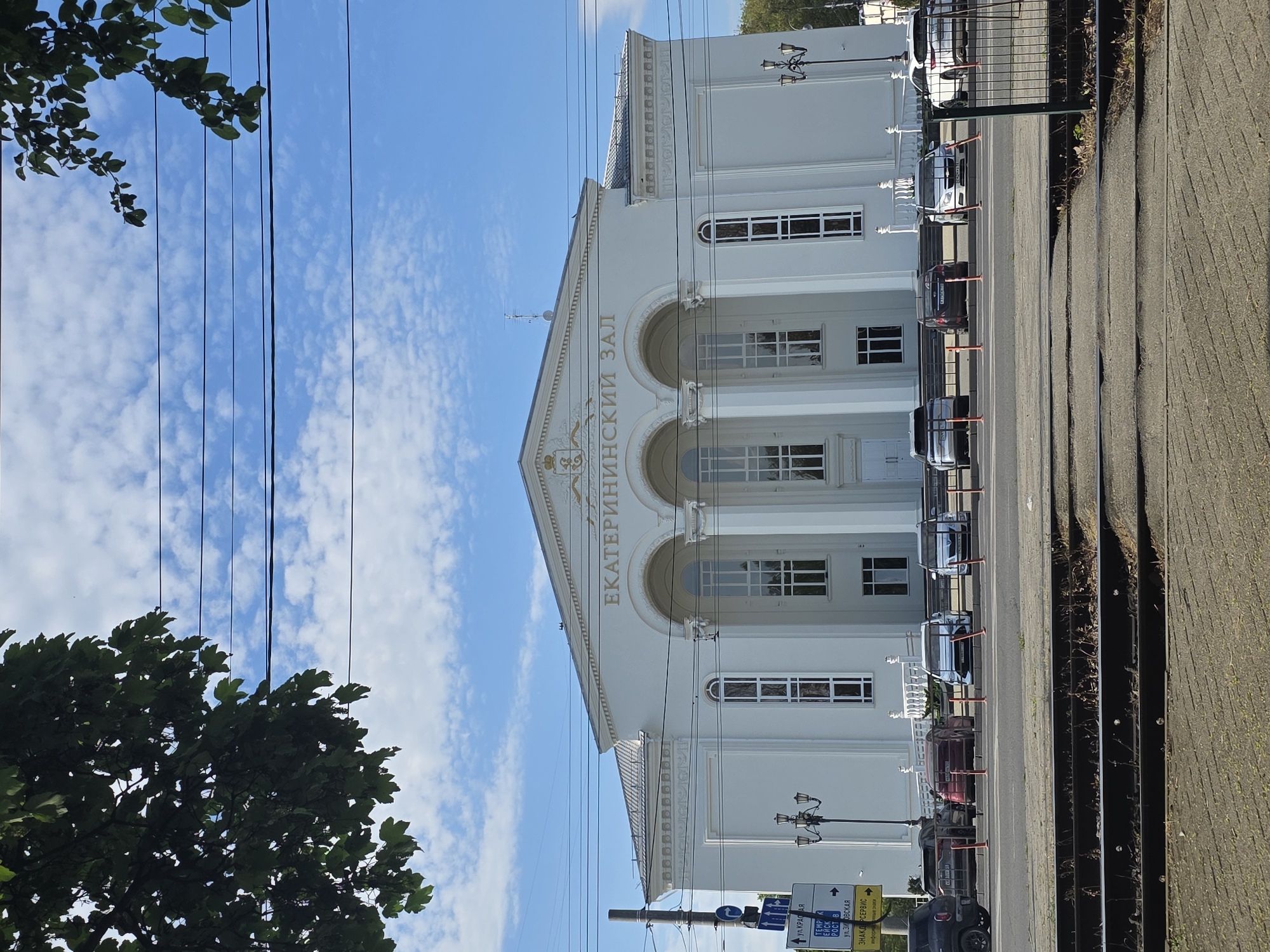 The image shows the facade of the Ekaterininsky Hall in Krasnodar, Russia. The building is a light gray color, with a classical architectural style, featuring columns, arched windows, and a triangular pediment. "ЕКАТЕРИНИНСКИЙ ЗАЛ" is inscribed in gold lettering on the pediment. Several parked cars are visible along the street in front of the building. Street lamps and street signs are also present. The sky above is blue with scattered clouds. Trees are seen on the left side of the frame.