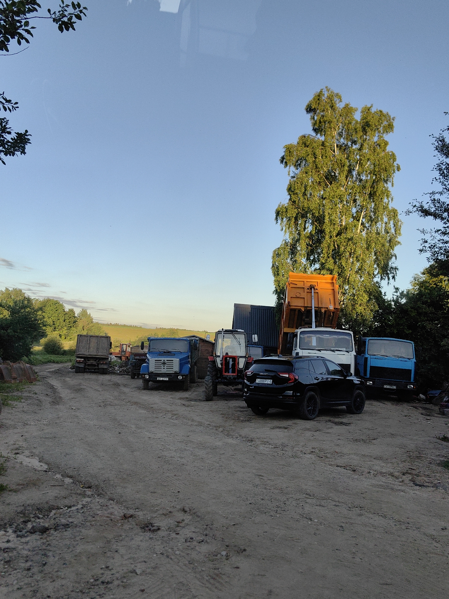 The image depicts a dirt road in a rural area, likely part of Дзямідавіцкі сельскі Савет, Belarus. The scene is dominated by several parked vehicles, including trucks and tractors. A large, orange dump truck is prominent, its bed raised. Beside it are two blue trucks, one of which has a license plate visible. A black SUV is parked in the foreground. In the background, trees and fields are visible under a clear blue sky. The lighting suggests it is late afternoon or early evening. The overall impression is one of a working yard or storage area for agricultural or construction equipment.