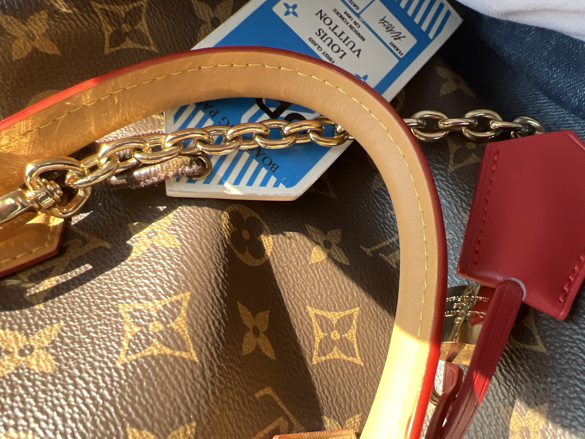 The image shows a close-up of a Louis Vuitton handbag, featuring the iconic monogram print. A light brown leather strap with red trim is visible, along with a gold chain. A blue and white boarding pass-style tag is attached to the chain, bearing the text "FIRST CLASS LOUIS VUITTON MAISON FONDÉE EN 1854". A red leather luggage tag is also visible. The handbag is resting on a surface that appears to be denim or a similar fabric. The lighting suggests an outdoor environment with sunlight casting shadows. This scene has no specific location in Letterkenny, Ireland.