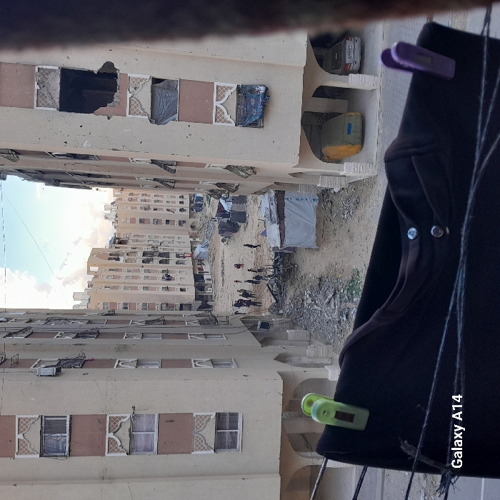 The image, taken from an elevated perspective, depicts a severely damaged urban residential area in Khan Yunis, Palestinian Territory. Multiple multi-story apartment buildings with light-colored facades and decorative window accents frame a central, unpaved area. Many windows on these buildings are broken, missing glass, or covered with makeshift materials; significant structural damage, including large open holes in upper floor walls, is evident on the rightmost building.
The ground level between the buildings is heavily covered with rubble, debris, and scattered refuse. Several makeshift tents or shelters constructed from tarpaulins are present in this central area. Approximately 10-15 individuals, consisting of adults and some children, are observed standing or walking in small groups amidst the debris.
In the immediate foreground, a dark garment hangs on a clothesline, secured by one green and one purple clothespin. Under the arched ground-floor supports of the building on the right, a vehicle is covered with a yellowish-green tarp, and a silver sedan is partially visible nearby. The sky above is partly cloudy.