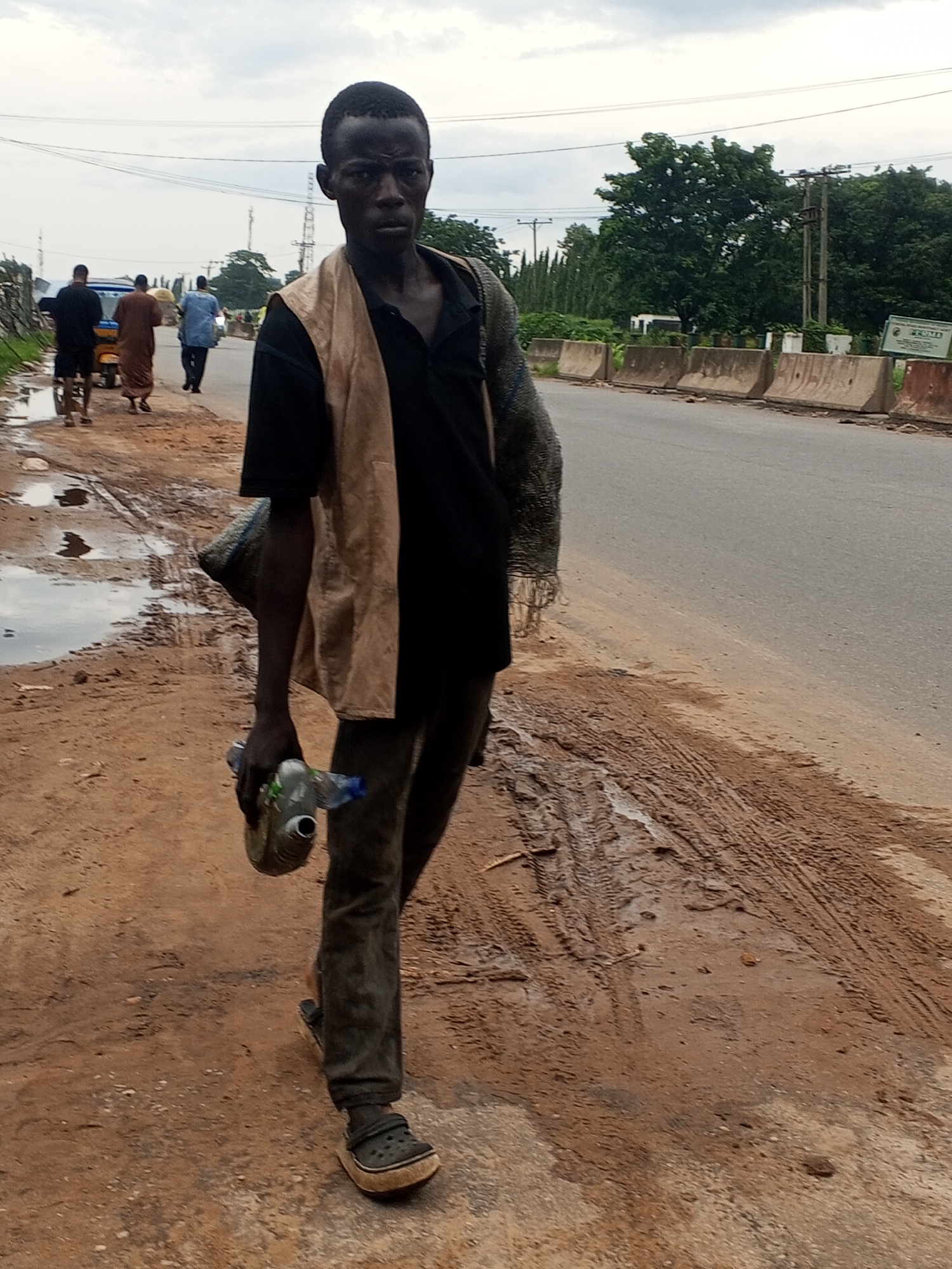 A man walks along the side of a road in Lokoja, Nigeria. He is carrying a grey bag on his left shoulder and a plastic bottle in his right hand. He is wearing a black shirt, a tan vest, dark pants, and black clogs. In the background, several other people walk on the road, and a yellow tricycle is visible. The road is paved, and the ground next to it is dirt and mud with tire tracks. Trees and utility poles are present in the distance. The sky is overcast.