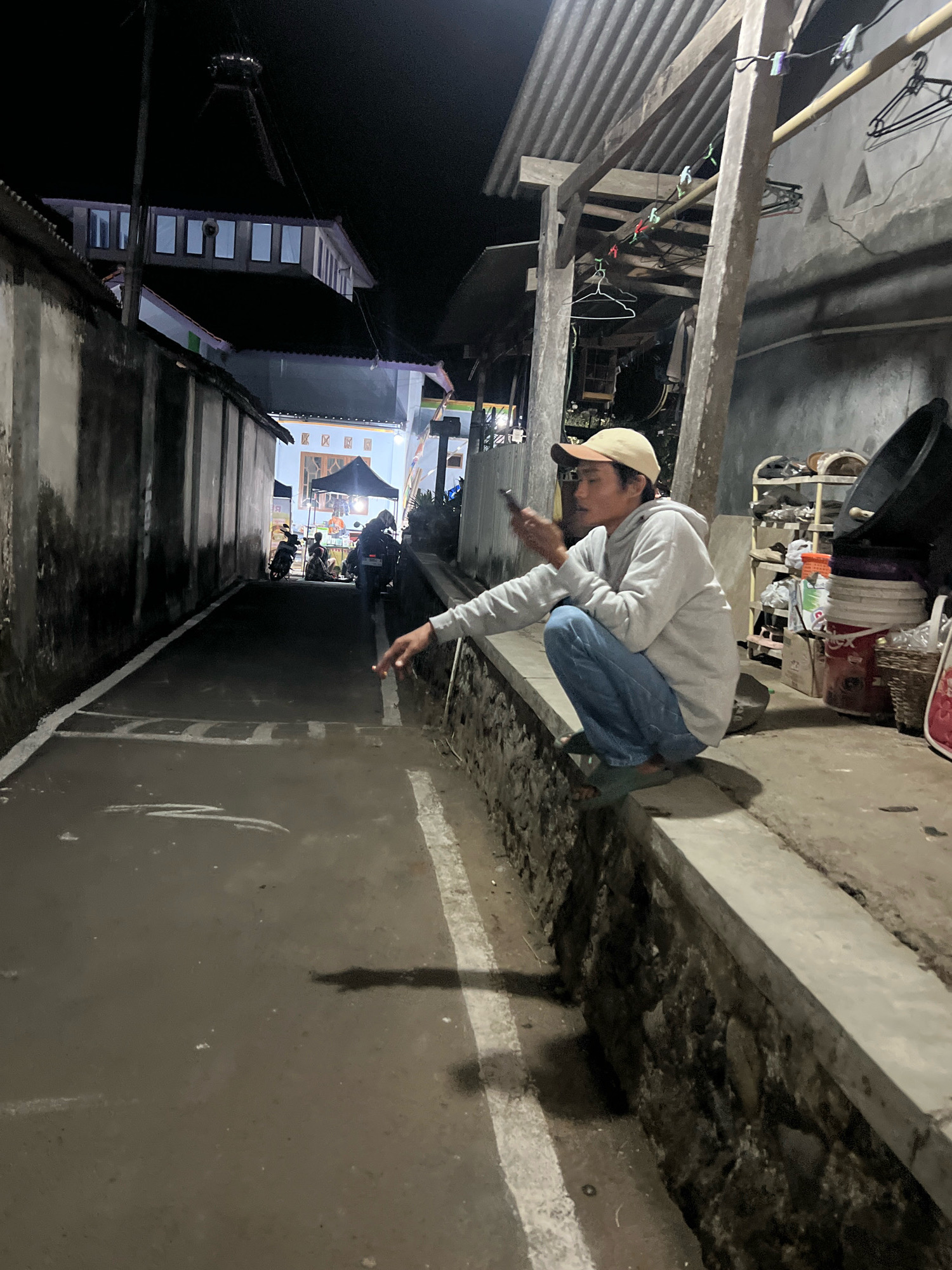 In Banjarsari, Indonesia, at night, a young man wearing a light-colored hoodie, blue jeans, a light-colored baseball cap, and green sandals crouches on a stone ledge. He is holding a dark-colored smartphone in his right hand, which is extended outward. His left hand is also extended, and appears to be holding a cigarette. The man is looking down and to his right. The background shows a narrow, dark alleyway with buildings on either side. The alleyway is wet and reflects the ambient light. There are some motorbikes parked in the distance, and a small market stall with people gathered around it. A corrugated metal roof structure is visible above the man, supported by wooden beams. There are clothes hanging from a line under the roof. Shelving units filled with various items, including containers and bags, are located to the man's right.