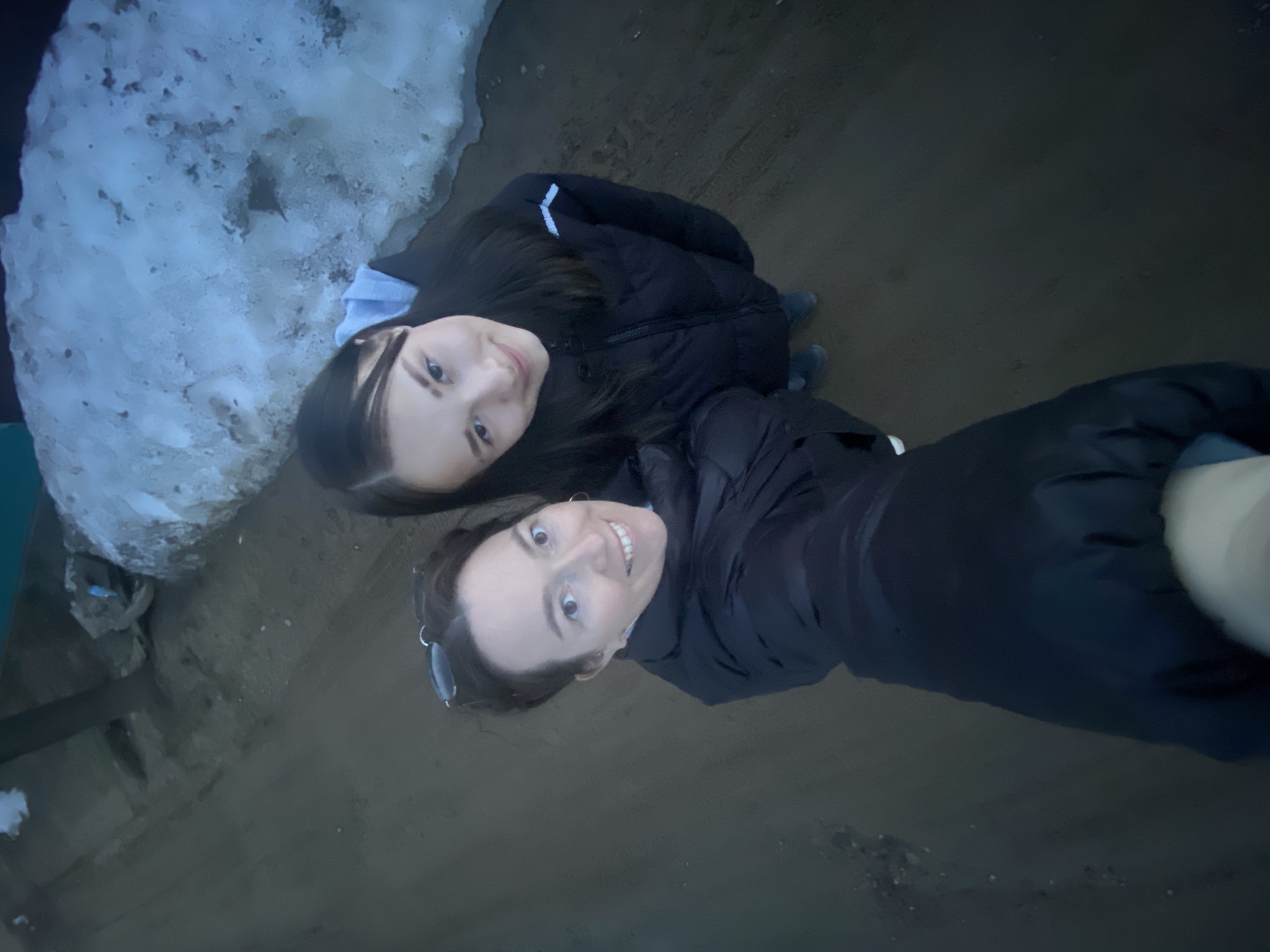 Two women are posing in a selfie taken from a high angle, with one woman slightly behind and to the left of the other. Both are wearing dark jackets, and the woman in the foreground has sunglasses perched on her head. To their left, there is a patch of snow. The ground appears to be a dark, muddy surface. This scene is located in Ольховка, Russia.