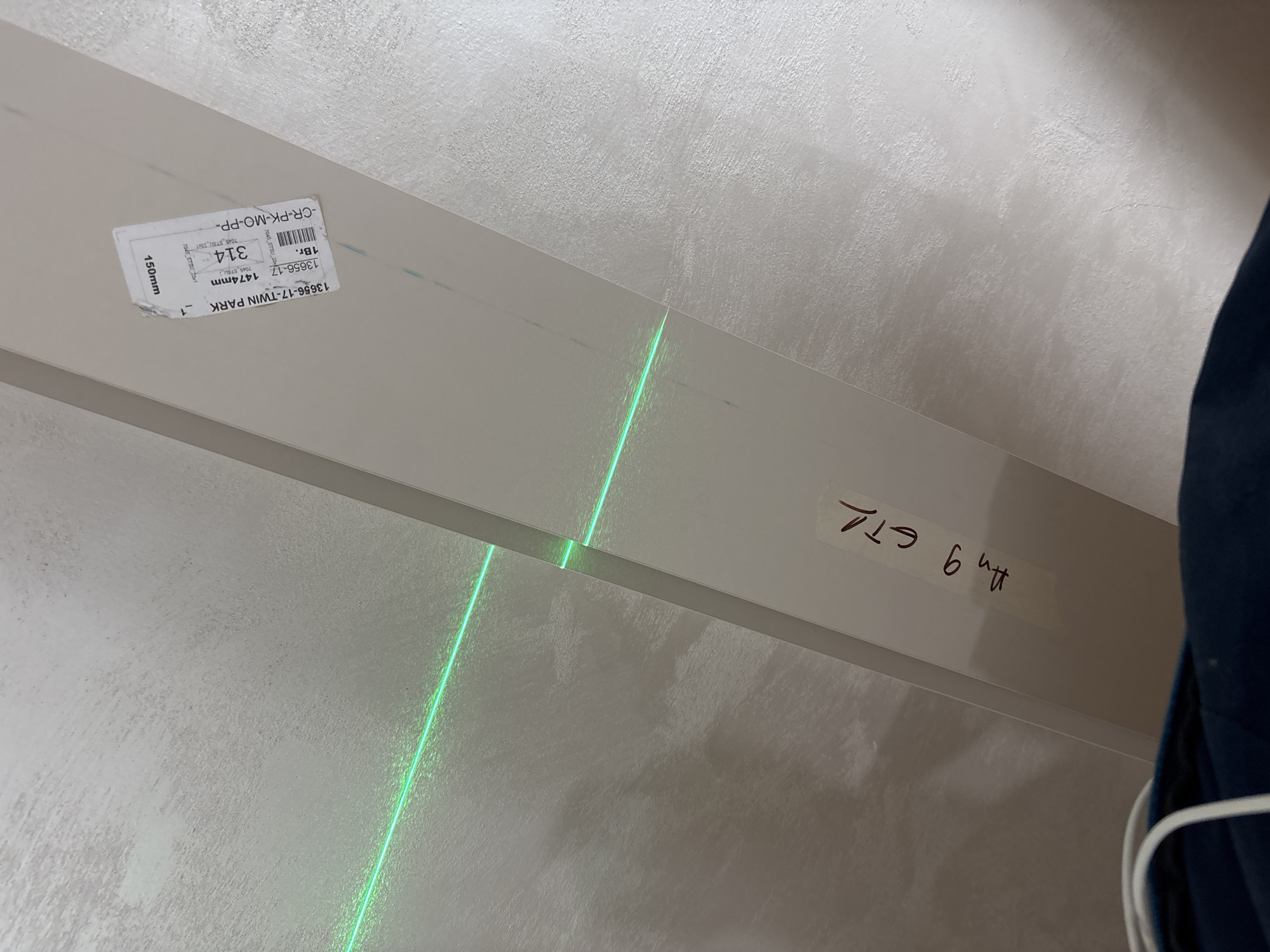 The image shows an angled view of a white beam, possibly a piece of trim or molding, with a bright green laser line crossing it diagonally. The beam has a label with product information, including the text "13656-17-TWIN PARK" and numerical details like "1474mm" and "314". There is also handwritten text on the beam, “Am 9 ET1”. The background appears to be a textured white wall. A portion of a person's dark blue clothing is visible in the right portion of the image. The location is not identifiable from the image.