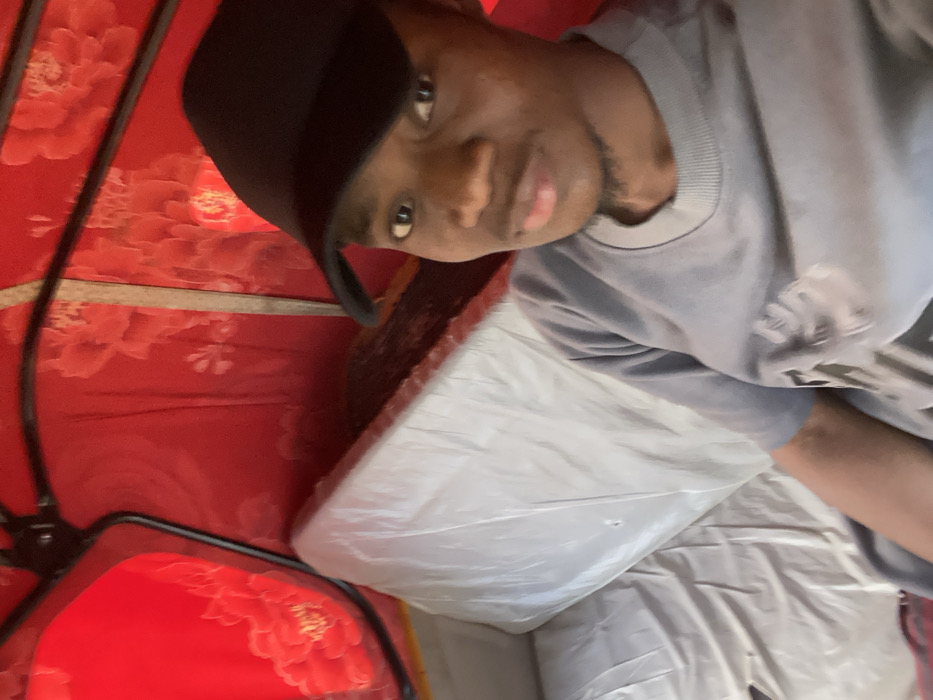 A single Black male is seated within the passenger compartment of a motorized tricycle (keke napep) in Minna, Nigeria. The individual wears a black baseball cap and a grey short-sleeved shirt, looking directly towards the viewer. The vehicle's interior is lined with red fabric featuring a lighter floral pattern. A white, plastic-covered seat is positioned to the left. Black metal frame elements are visible against the red interior.