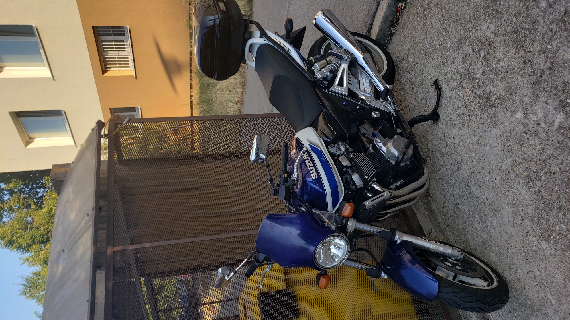 A blue and white Suzuki GSX 1400 motorcycle is parked on a concrete surface next to a yellow dumpster and a brown metal fence. The motorcycle has a dark-colored top box on the rear seat. In the background, a light-colored building with brown accents is visible, along with trees and a clear blue sky. The scene appears to be an outdoor urban setting.
