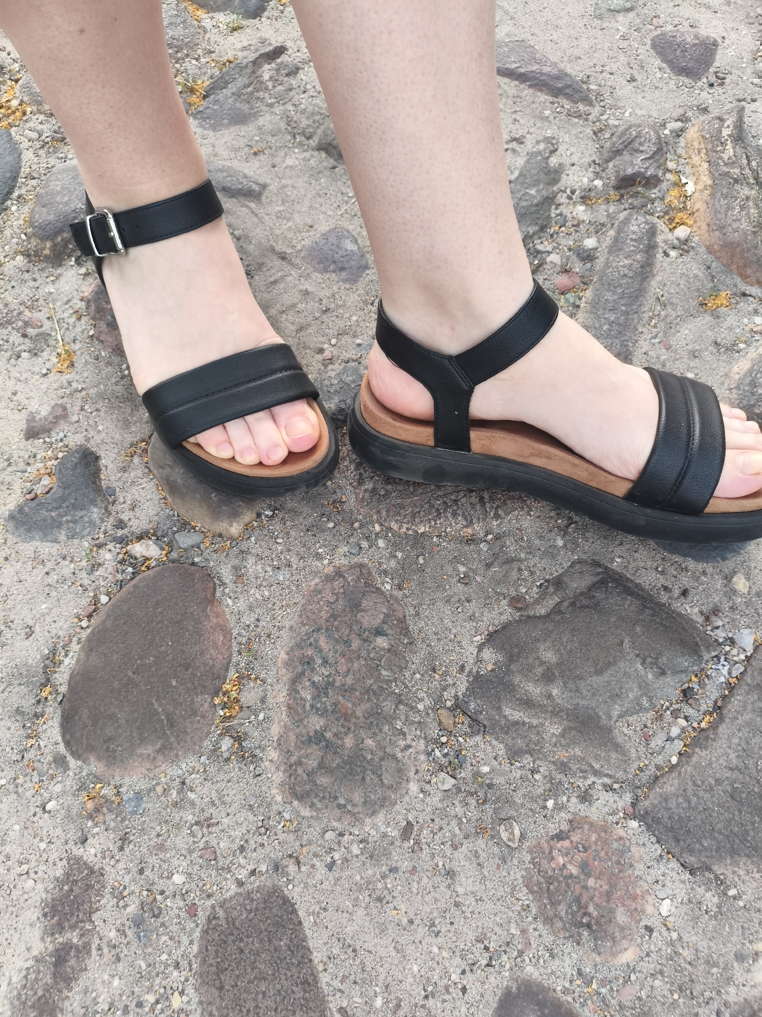 The image shows a close-up, low-angle shot of two bare feet wearing black sandals. The sandals have a thick sole and an ankle strap with a buckle. The feet are standing on a cobblestone surface, with some small pebbles and yellow debris scattered around. The lighting suggests it is daytime. The specific location is the cobblestone pathway in front of the Modlin Fortress in Nowy Dwór Mazowiecki, Poland.