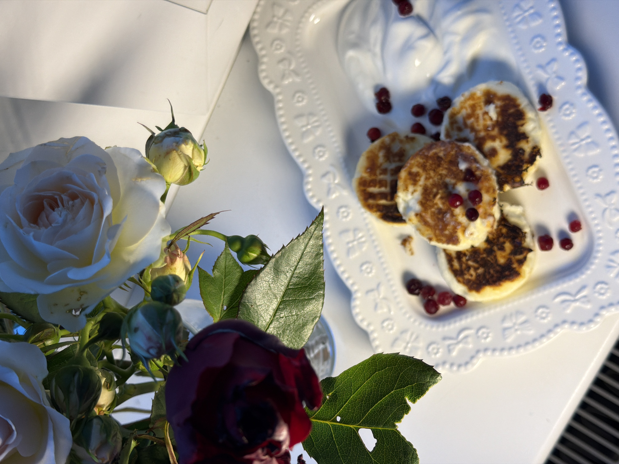 The image shows a still life arrangement. In the foreground, there is a bouquet of roses, including white and dark red blooms. The roses are in a vase (partially visible). Adjacent to the flowers is a white, decorative plate containing several small, round pancakes or fritters topped with red berries. The plate also has a molded design of cherubs. Scattered around the pancakes are more of the same red berries. The scene is set on a white surface, possibly a table, with a blurred striped pattern visible near the bottom right corner of the image. A white box or frame is present in the upper left corner. It's difficult to pinpoint the precise location in Rossiyskiy, Russia, without further context or landmarks in the image.