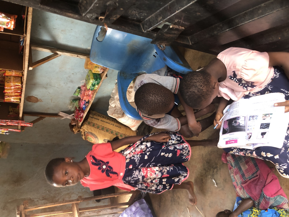 The scene depicts an interior space in Ukwu-Abwa, Nigeria, functioning as a small, rudimentary shop or dwelling. Four children are present. A young girl, barefoot and wearing a coral top with a floral skirt, stands centrally, facing slightly towards the viewer. To her right, two children are seated on the concrete floor; one, in a grey and white striped shirt, leans forward, while a girl in a light pink top and floral skirt holds an open textbook containing images and text, actively writing with a pen. A fourth child is partially visible lying on a plaid blanket in the lower-left corner. The light blue walls are unpainted and show wear. Rustic wooden shelves, supported by planks, are mounted on the back wall, displaying various packaged goods such as sweets, snacks, and sachets. Large woven sacks are stacked on the floor beneath the shelves. An overturned blue plastic chair is visible on the right, near a dark, possibly metal, frame or door structure. A small, pale lizard is on the floor near the feet of the standing girl.