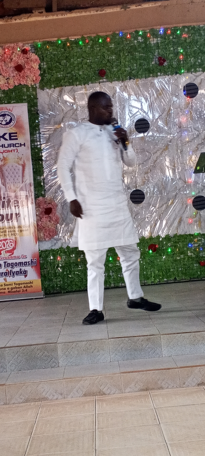 The image displays an indoor scene within a building in Akwanga, Nigeria, showing a male individual on a raised platform. The man, an adult with dark skin and short dark hair, is positioned slightly right of the frame's center. He wears a white long-sleeved tunic, white trousers, and black shoes. A gold-colored ring is visible on his right index finger. He holds a blue and black microphone to his mouth with his right hand, his left foot slightly raised, suggesting movement.
The background consists of layered decorations. The uppermost section features a horizontal panel of artificial green foliage, interspersed with small red, green, and blue LED lights. Below this, a silver-colored, crinkled, reflective material forms a backdrop, on which several circular black objects with horizontal stripes are affixed. Clusters of artificial pink and red flowers are placed on the green foliage panel and the silver backdrop.
To the left, a tall, vertical banner is visible, displaying a "BAPTIST CONVENTION" logo, "KE CHURCH (LIGHT)", an illustration of a white armchair, and the text "UNLIMITED FAVOUR 2023". Further text includes "Za Sami Tagomashi" and a Bible verse reference "Prov 3:1 NLT". The man stands on a light-colored tiled platform, which transitions via a step to a lower, similarly tiled floor in the foreground. The top edge of the image reveals a brown structural element, possibly a ceiling beam.