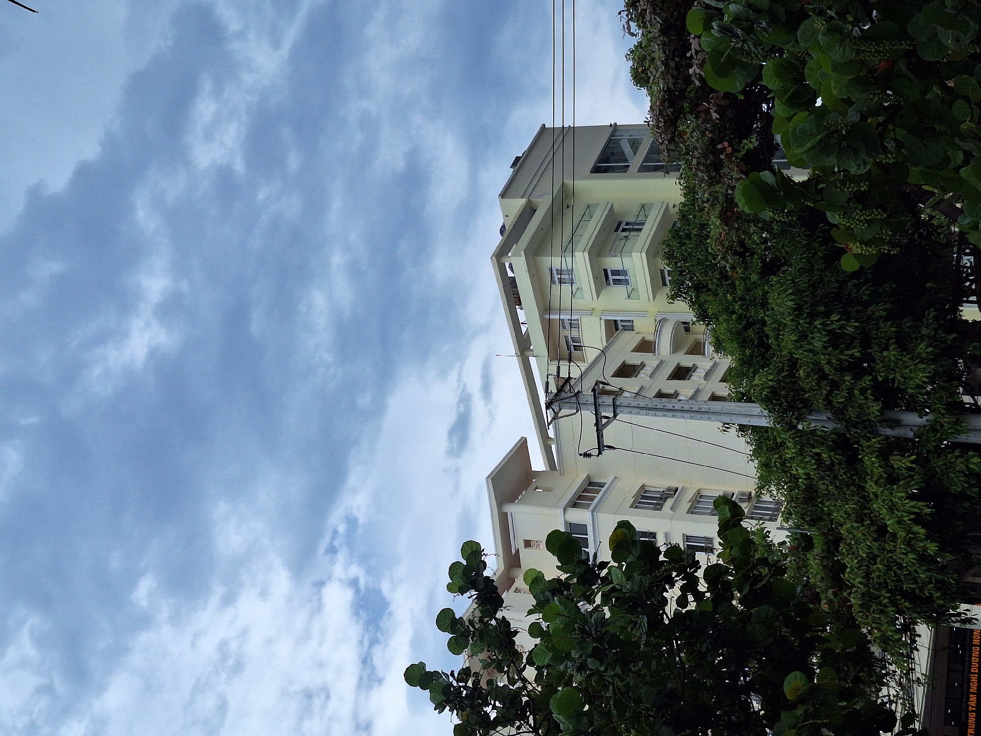 The image shows a multi-story, light yellow building with balconies and large windows, partially obscured by lush green trees and foliage in the foreground. A utility pole with electrical wires is visible to the right of the building. The sky above is overcast with gray clouds. A sign is partially visible at the bottom left of the image, appearing to be in Vietnamese. The building's architectural style and the presence of tropical vegetation suggest a location in a coastal city. This scene is consistent with the environment found in Nha Trang, Vietnam, a well-known beach resort city.