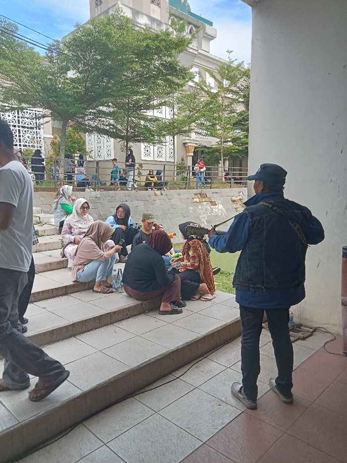 The image captures an outdoor gathering of people in Singaparna, Indonesia, during daytime, under what appears to be an overcast sky.
In the foreground, a man with his back to the viewer, wearing a cap, a blue long-sleeved shirt, and a dark vest, stands near a white wall. He is holding and seemingly playing a stringed instrument, possibly a rebab or a fiddle. On the tiled steps to his left, several individuals are seated, mostly women, some wearing hijabs. They appear to be casually socializing, with a few looking towards the man or at objects in their hands. Bottles and bags are visible near them. On the far left, a person wearing a white t-shirt and grey pants walks past, partially visible.
The scene extends into the mid-ground, where more people are visible near a low stone wall and a metal railing. Some are sitting on benches, while others are standing or walking. In the background, a large, ornate building with a prominent tower, resembling a mosque, stands amidst lush green trees. Its light-colored facade features intricate window designs. A grassy area is visible to the right of the steps.
The overall atmosphere is relaxed and social, suggesting a community gathering or a public event near a mosque. No discernible text is visible in the image.