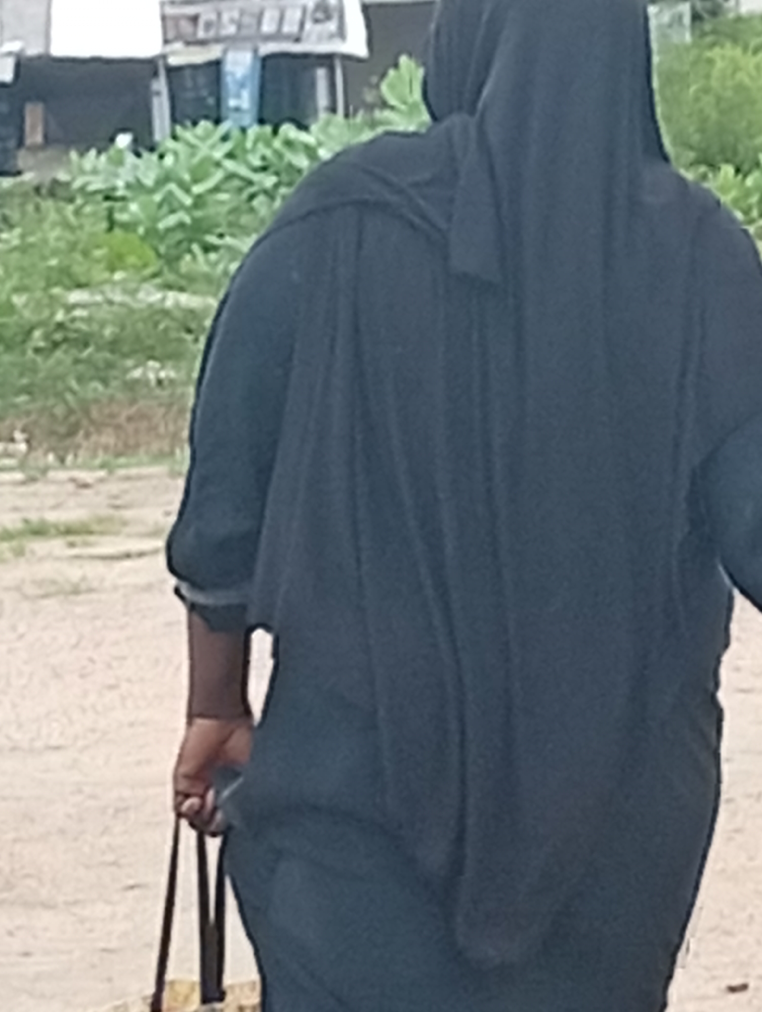 The image depicts a person from behind, wearing a black hijab and abaya, walking away from the camera. The person is carrying a dark-colored bag with two straps in their left hand. The background shows a sandy or dirt path with some vegetation and what appears to be a building with a white roof and some structures behind it. The environment suggests a semi-urban or rural setting. The overall lighting indicates it is daytime. The person's skin tone is dark. The image quality is low, with visible pixelation.