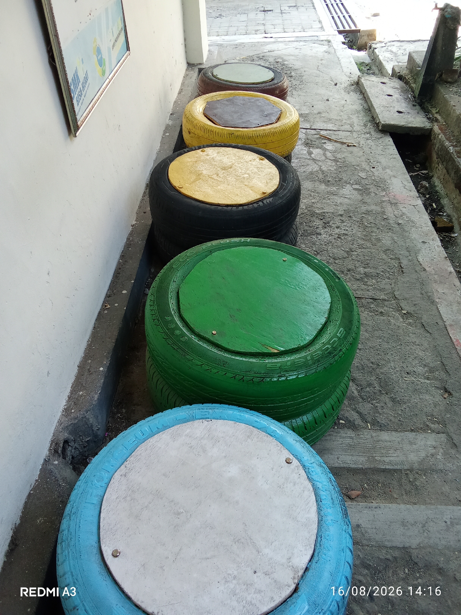The image shows a row of repurposed tires painted in different colors, arranged along a concrete pathway next to a white wall. The tires are stacked and topped with wooden seats, creating a makeshift seating area. From foreground to background, the colors of the tires are blue, green, yellow, and brown. The environment appears to be outdoors, possibly in a public space within the city of Surakarta, Indonesia, as indicated by the timestamp overlay. The pathway is made of concrete slabs, and a drainage ditch is visible to the right of the seating area. A framed poster is affixed to the white wall. The scene is well-lit by natural daylight.
