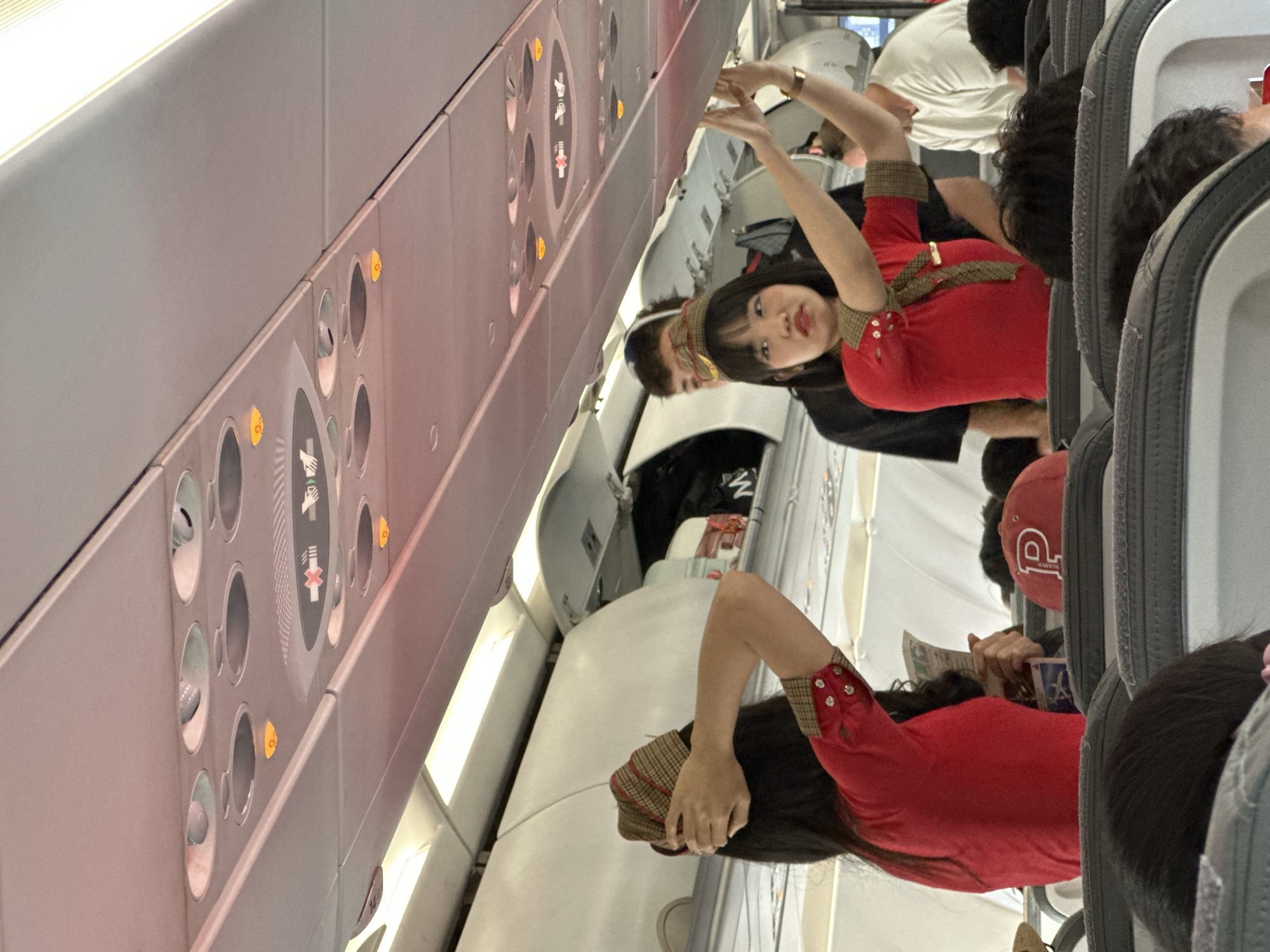 In an airplane cabin, a flight attendant in a red uniform with a checkered collar and buttons on the sleeves is the primary subject. She is gesturing with both hands, her arms raised, and her face is turned towards the camera with a slight smile. Her dark hair frames her face. She is wearing a checkered headband.
Behind her, other passengers are visible. One man, wearing a white t-shirt, is seen in the background. Another passenger in the foreground, also in a red uniform similar to the flight attendant's, is visible from the waist down, holding what appears to be a brochure or pamphlet in their hand. Other passengers are seated in the rows of airplane seats, some with dark hair. One passenger in the foreground is wearing a red baseball cap with a white logo.
The scene is set within an airplane, indicated by the rows of seats, overhead compartments with air vents and lights, and the general interior of the cabin. The overhead lights are on, suggesting it is likely daytime or that the cabin is artificially lit. The image has a slightly blurred quality, and the perspective is from a low angle, looking up towards the flight attendant. The overall atmosphere seems casual and potentially posed, given the flight attendant's direct gaze and gesture towards the camera. There is no visible weather information. The location cue is Kuta, Indonesia.