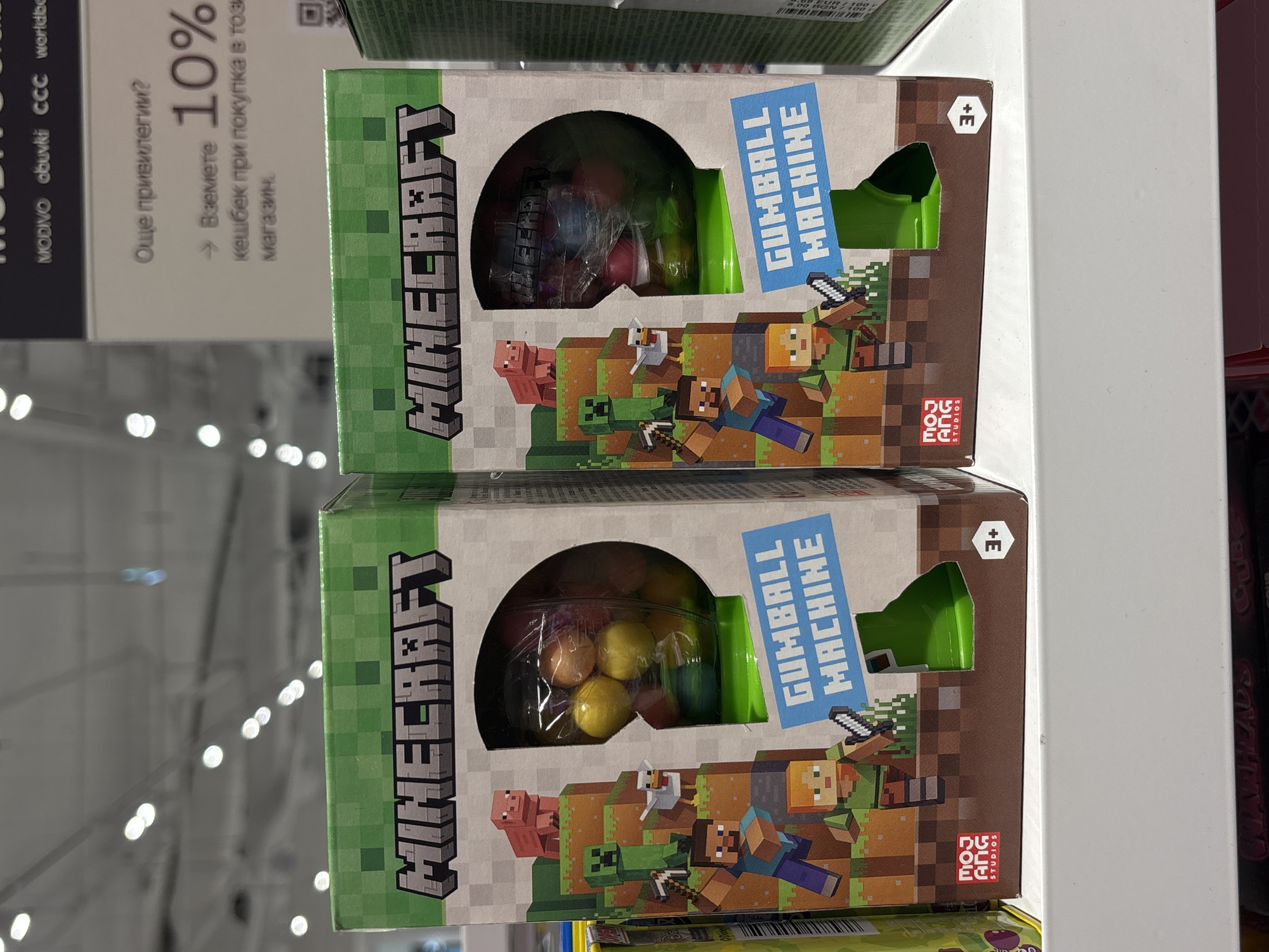 The image shows two boxes of "Minecraft Gumball Machine" toys stacked on a shelf. The boxes feature the Minecraft logo and imagery. The product appears to be a gumball machine. The background is blurred, suggesting the location is a store. Text in Bulgarian on a nearby sign ("Още привилегии? → Вземете 10% кешбек при покупка в този магазин.") indicates the store is located in Bulgaria. The MODIVO, obuvki, CCC, and worldbe logos also appear. Given the presence of Bulgarian text, the location is possibly in Varna, Bulgaria.