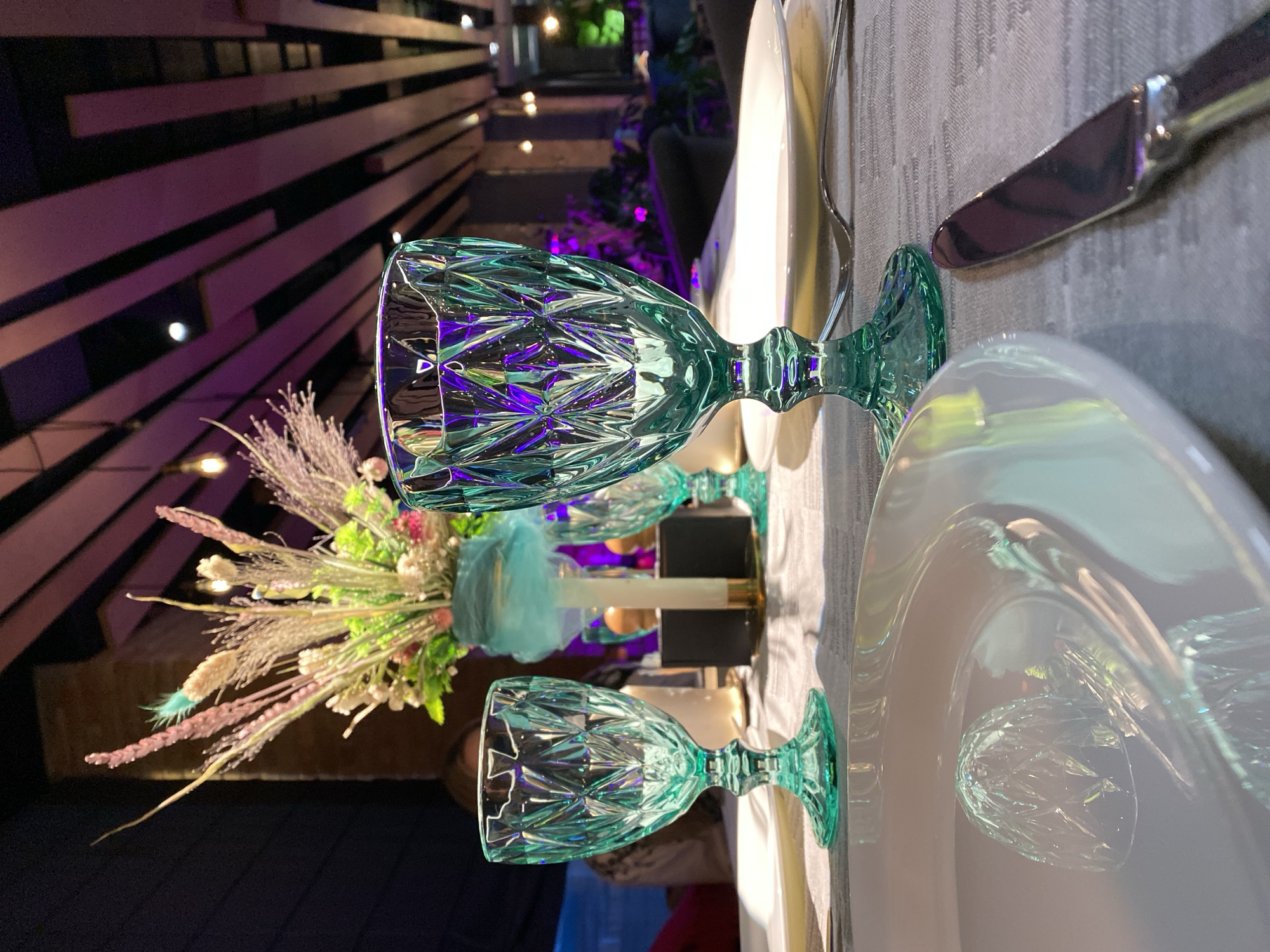 The image depicts a beautifully set table in what appears to be a restaurant. Two stemmed wine glasses, made of faceted teal glass, are prominently featured. The glass on the right is in the foreground and is filled with light that refracts through its intricate design, showing hues of green and purple. A white plate is positioned in front of this glass, and a knife rests to the right of the plate. In the background, a floral arrangement with dried grasses and greenery sits in a teal vase, with a white candle behind it. The ceiling features modern wooden slats with purple and white lighting, suggesting a contemporary ambiance. A person is partially visible in the blurred background. The setting is dark, with ambient lighting creating a mood for dining. There is no indication of the city of Myltsi, Ukraine, or any specific location.