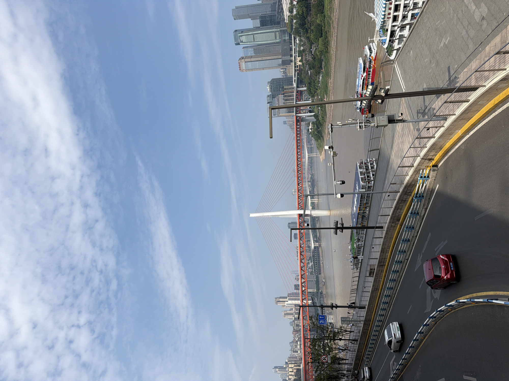 This is a full shot of a sunny day in Yuzhong District, China, with a focus on a modern cityscape and a wide river. The sky is blue with scattered white clouds. In the foreground, a multi-lane road curves to the left, with a red car and a white car driving on it. A yellow and blue striped barrier runs along the side of the road.
Beyond the road, a paved walkway lines the riverbank, separated by a metal railing. Several people are visible walking along the walkway. A large, multi-story passenger boat is docked at the walkway, and another boat with a striped awning is partially visible.
The river is wide and brown, with some smaller boats on its surface. A prominent orange suspension bridge spans the river in the middle ground, supported by a tall white pylon. The city skyline rises behind the bridge, characterized by numerous modern high-rise buildings. Trees and vegetation are visible on the riverbanks. The overall scene depicts a bustling urban environment with a blend of infrastructure and natural elements.