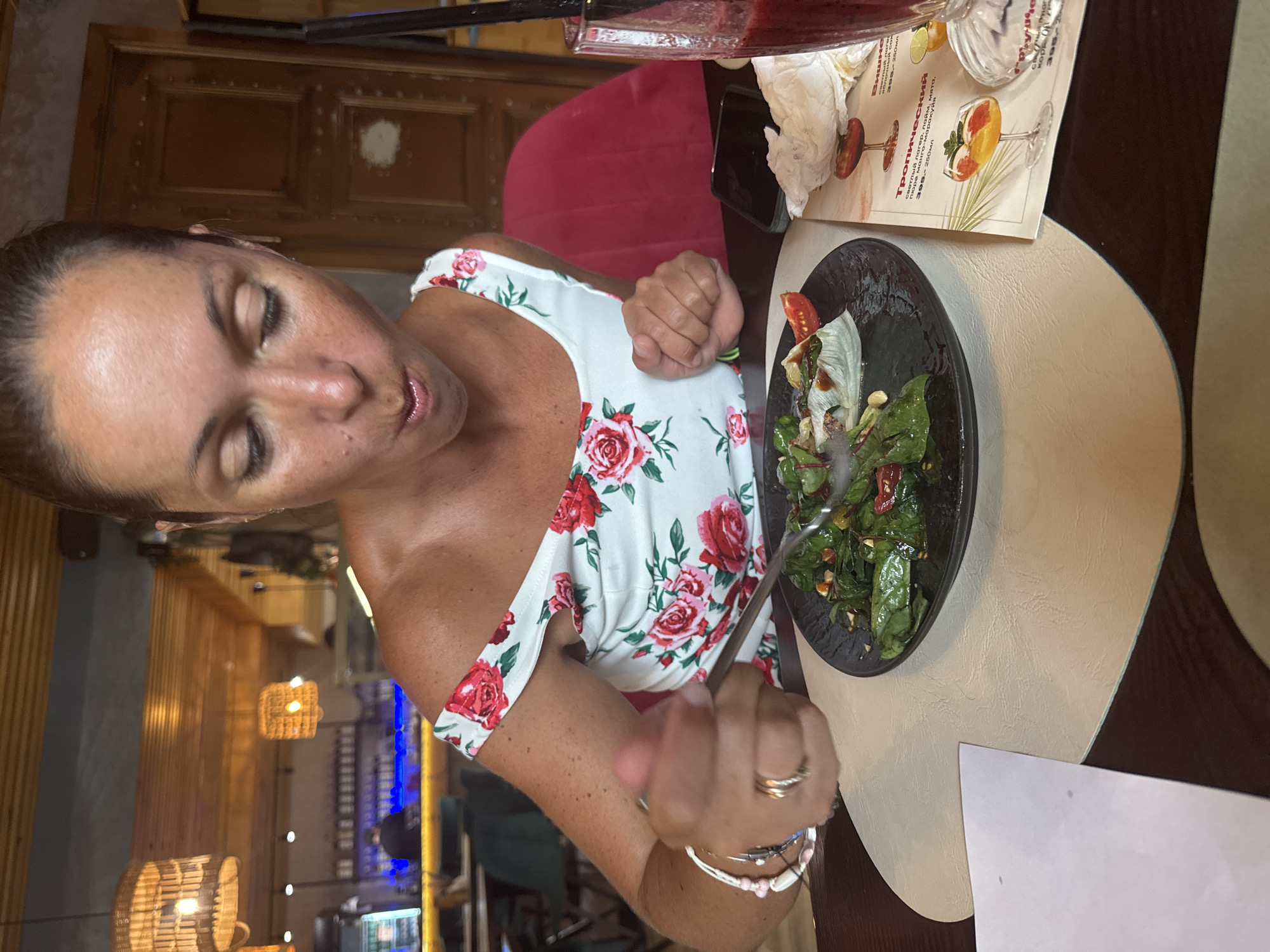 A medium shot captures a woman dining in a dimly lit restaurant. She is wearing a white dress with a red floral pattern and is leaning forward, eating from a black plate with a fork. In front of her is a glass of red juice with a straw. The background shows wooden ceiling beams, hanging lights, and a bar area with bottles. A menu with images of cocktails is visible on the table. The overall impression is of a relaxed meal in an indoor establishment.