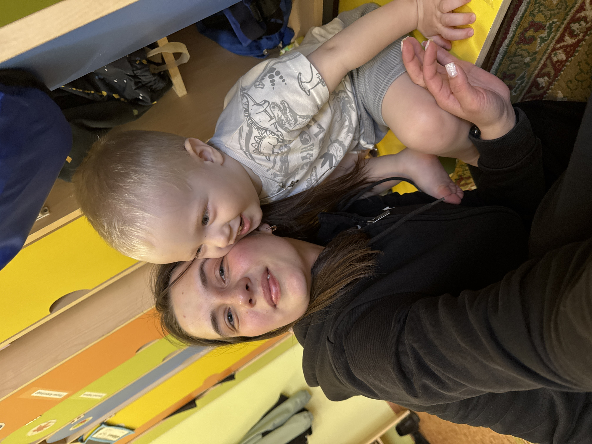 This image, taken in Chaikovsky, Russia, depicts a heartwarming moment between a young adult female and a toddler, both smiling warmly. The image is oriented sideways, suggesting the subjects might be lying down or in a relaxed pose.
The female, with brown hair, is wearing a black hooded sweatshirt. Her left hand is visible, showing a white, glittery nail polish on her index finger. She has a gentle, happy expression as she looks towards the camera.
The toddler, with light blonde hair, is nestled close to the female, his cheek pressed against hers. He is also smiling, his bright eyes wide open. He is dressed in a light-colored t-shirt featuring grey cartoon dinosaur graphics and grey shorts. His bare feet are visible, one resting on the female's arm, emphasizing their close physical bond.
The background suggests an indoor setting, likely a child's play area or room. Colorful wooden furniture, possibly a shelving unit or drawers, is prominent, with segments in yellow, orange, blue, and natural wood tones. Faint, unreadable text labels appear on some of these colorful sections. A patterned rug with traditional motifs in shades of green, red, and yellow is visible on the right. Other miscellaneous items, including a dark blue fabric object, are partially visible in the periphery. The overall scene conveys a feeling of joy and affection. The time of day is not discernible.