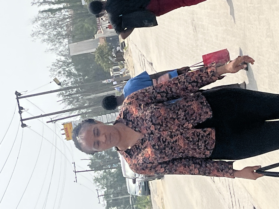The scene depicts a daytime urban street in Lekki, Nigeria. In the foreground, an adult female with dark, braided hair walks towards the viewer's left, looking slightly downward. She wears a long-sleeved collared shirt with a black base and orange/beige floral patterns, paired with black trousers. Her left hand holds a dark object, possibly a bag.
In the mid-ground, behind the primary subject, an adult male walks away from the viewer, attired in a blue t-shirt and light-colored trousers, carrying a red shopping bag in his right hand. Further to the right, another adult individual is partially visible, wearing a dark top and red pants, with their left hand positioned on their hip. A light-colored sedan is visible on the street to the left.
The environment features a light-colored paved road surface. Multiple wooden utility poles supporting numerous overhead power and communication lines dominate the mid-background. A prominent yellow sign with red "DHL" lettering is affixed to one of these utility poles, indicating proximity to a DHL service point. Another sign, partially visible as "GALLERY," is located further down the street to the right. Trees and vegetation line the background, and the sky is bright and overcast. The overall setting is a commercial or mixed-use street within Lekki, Nigeria.