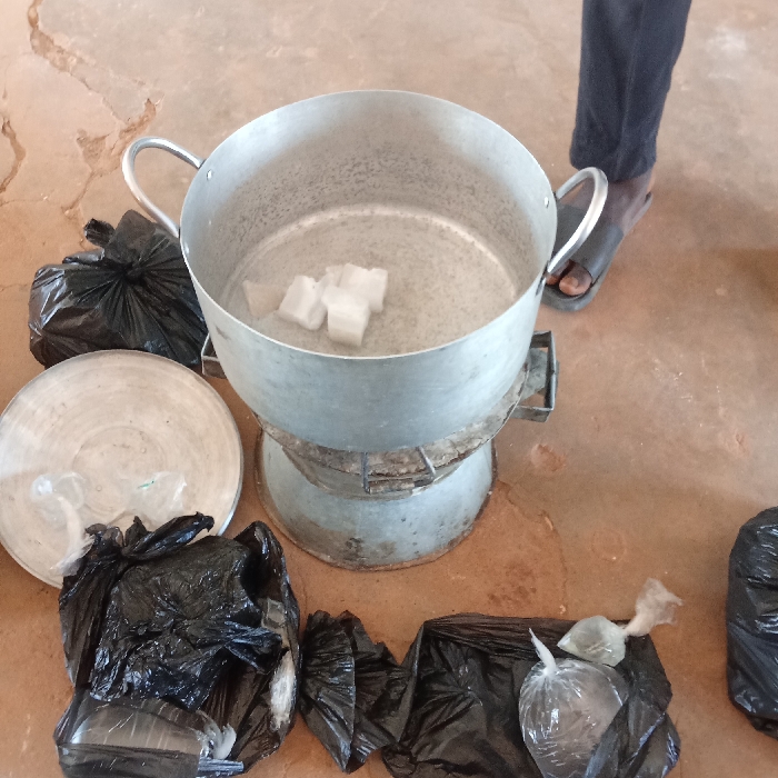 The scene in Akwanga, Nigeria, depicts an informal processing area on a reddish-brown, cracked floor. A large, dull silver-colored aluminum pot with two handles is positioned on a metallic charcoal stove. Inside the pot, approximately five irregularly shaped white solid blocks are partially submerged in liquid. To the left of the pot, a silver-colored metal basin or lid rests on the floor. Multiple black plastic bags are distributed around the central apparatus; some are tied shut, while others contain smaller, clear plastic pouches. Two clear pouches in the foreground contain a light-colored liquid or semi-solid substance and are knotted. The lower portion of a person's legs and feet, wearing black sandals, is visible to the immediate right of the pot.