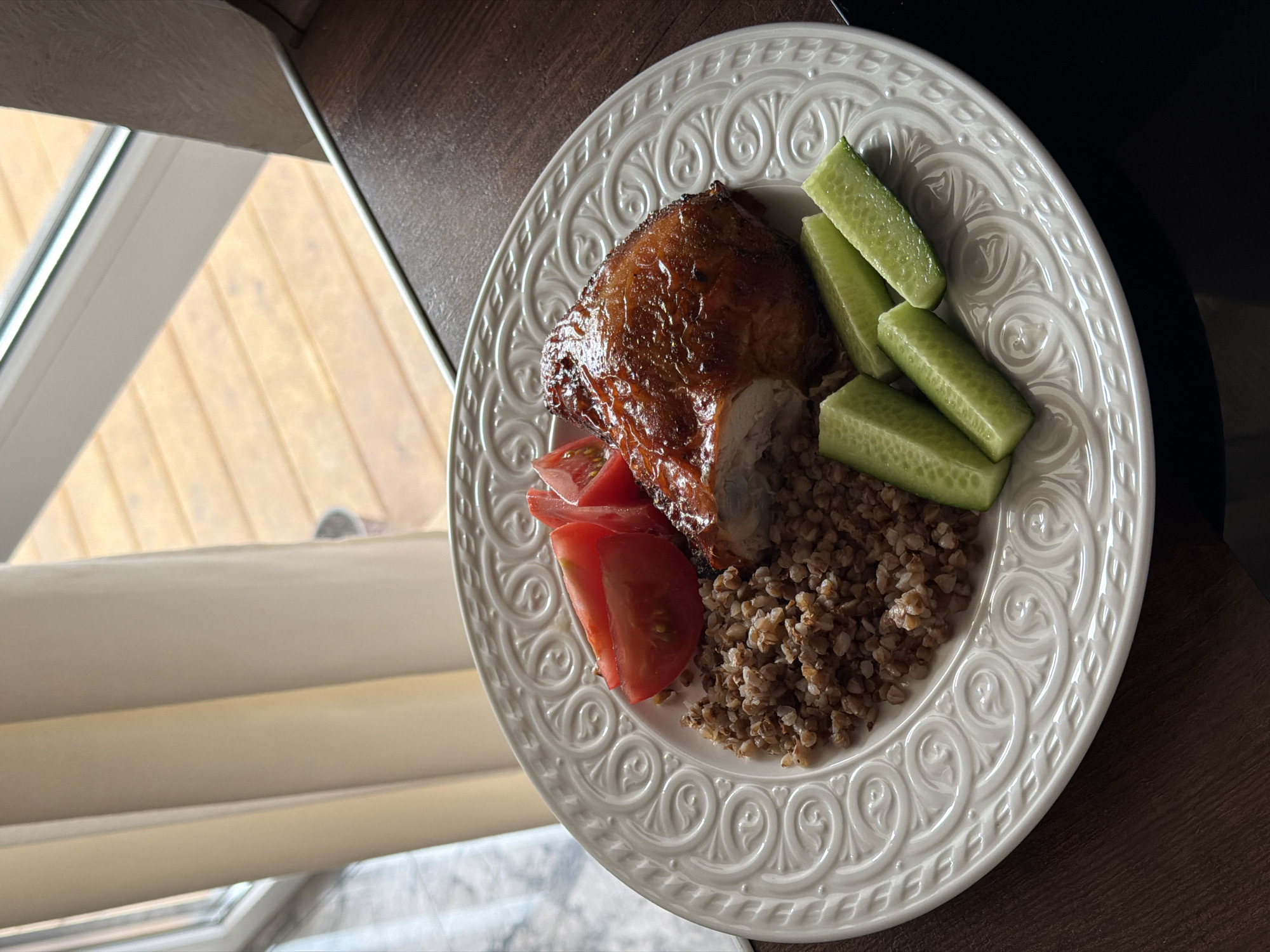 The photograph depicts a white plate containing a meal consisting of a piece of cooked chicken with a slice removed, buckwheat, sliced tomatoes, and cucumber sticks. The plate rests on a dark wooden surface near a window. The background visible through the window shows a wooden structure or fence. The image appears to have been taken indoors, likely in a dining or kitchen area. The plate itself is decorated with a pattern of repeating scroll designs. The location may be in Rossiyskiy, Russia, but there is no contextual evidence to confirm the city.