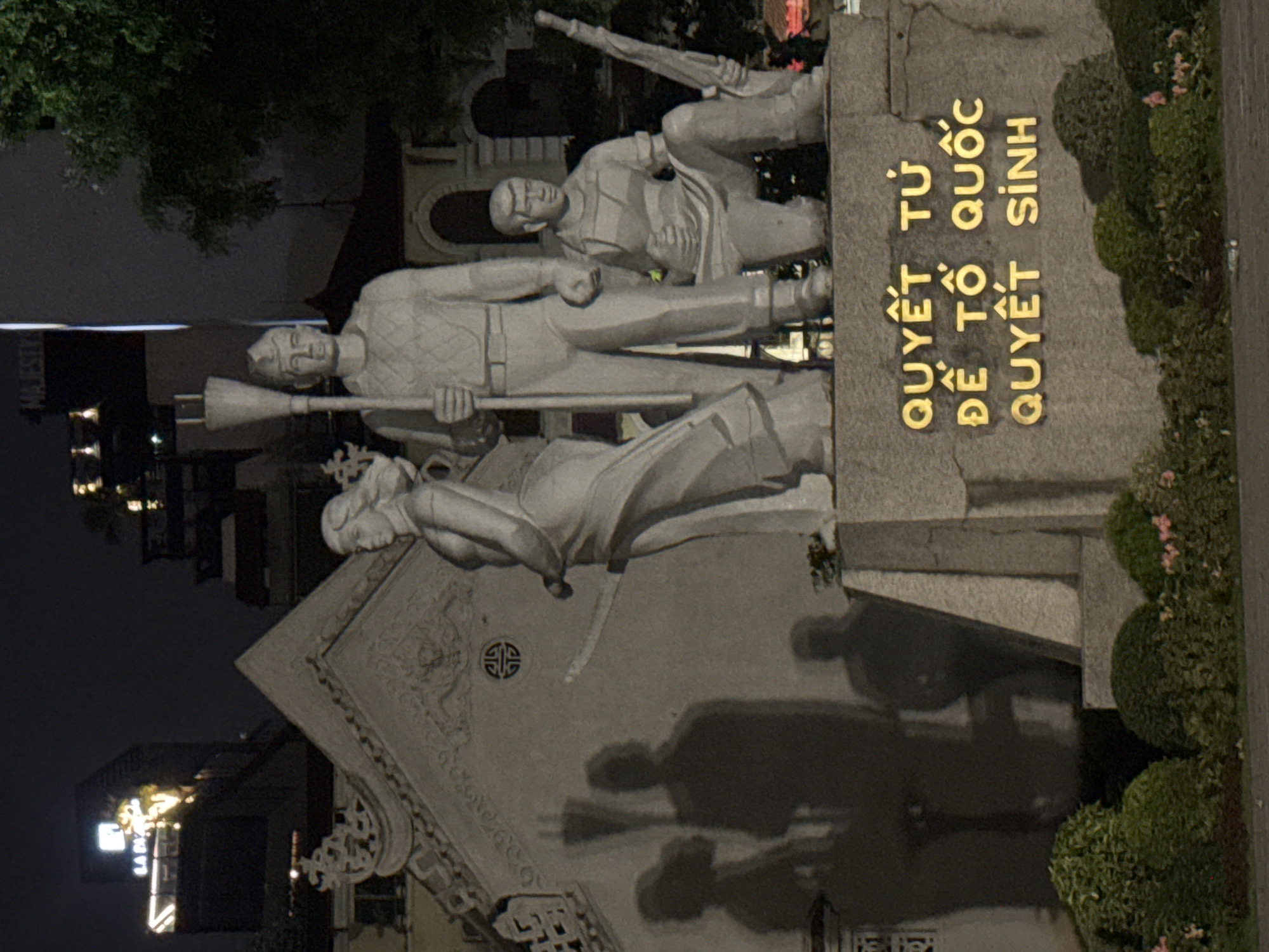 This is a nighttime photograph taken in Hanoi, Vietnam, showcasing a monument with several statues of people. The monument features a stone inscription with golden lettering. The statues depict individuals in what appears to be a historical or patriotic scene, possibly related to defense or struggle, given the presence of what looks like a rifle. One statue of a man holds a long, pole-like object, while another man behind him appears to be aiming a rifle. A female figure is positioned lower down, seemingly in a protective or supportive stance.
The setting is dimly lit, suggesting it is nighttime. The monument is situated outdoors, with greenery and bushes visible to the right. Shadows cast by the statues and possibly other elements are prominent on a textured, possibly carved, wall behind them. The overall mood is solemn and commemorative.
The visible text on the stone monument is in Vietnamese: "QUYẾT TỨ QUỐC ĐỂ TÔ QUỐC QUYẾT SINH". This translates to "Determination for the Nation, to sacrifice for the Nation." The golden lettering is clear and stands out against the stone. In the background, faint lights suggest the presence of buildings or streetlights, indicating an urban environment.