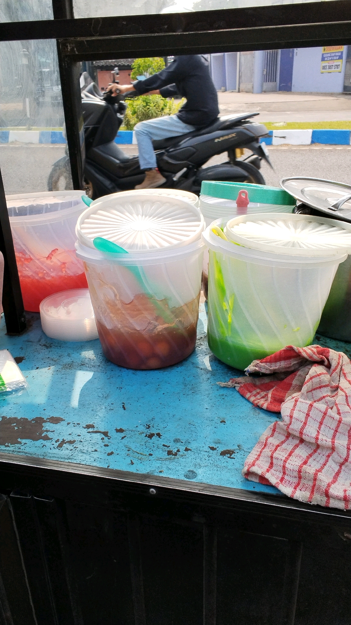 In the city of Banjaragung, Indonesia, on a bright day, a person rides a black motorcycle. The scene is viewed from behind a blue, worn tabletop. In the foreground, there are three large plastic containers filled with drinks. The container on the left contains a red liquid with pieces of fruit or jelly. The middle container holds a brown liquid with round, dark brown spheres, possibly fruit or candies, and a teal spoon. The container on the right contains a green liquid. Next to the containers is a white, circular object, possibly a lid or small bowl, and a red and white checkered cloth. A person is visible in the background, wearing a dark long-sleeved shirt and light blue pants, riding a motorcycle. The motorcycle has the word "NMAX" on its side. The environment appears to be outdoors, with a street and buildings visible in the distance.