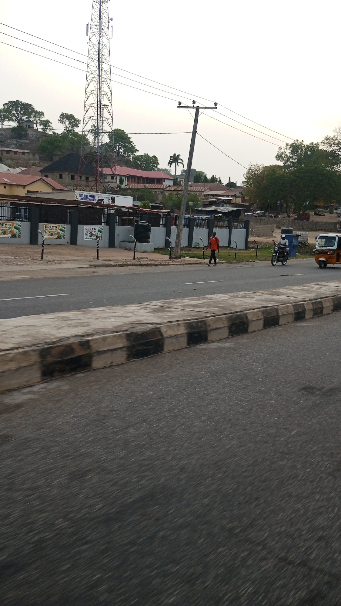 This image captures a street scene in Lokoja, Nigeria, likely in the late afternoon or early evening under a bright, slightly overcast sky. The foreground shows a multi-lane asphalt road with a concrete median featuring black and white striped curbs.
On the right side of the road, a person wearing an orange shirt walks along the shoulder. Further down the road, a motorcyclist in a white helmet is visible, and a yellow and blue tricycle (known as Keke Napep in Nigeria) is also present.
Beyond the road and median, there's a dark fence with multiple advertising boards. Text visible on these boards includes "DREY'S" (repeated) and "DAYLIGHT PERFECT LOGICS INVESTMENT LTD". Behind the fence, several buildings are visible, including what appears to be a commercial or residential structure with a black water tank placed on a stand. White-and-black striped street light poles are also spaced along the roadside.
In the mid-ground, a tall communication tower stands prominently, with power lines running across the frame, supported by a wooden utility pole. The background features rolling hills covered in trees and scattered buildings, some with red roofs, adding to the urban-rural blend of the landscape. A distinct palm tree is also visible against the sky. The overall impression is that of a typical street environment in a Nigerian town.