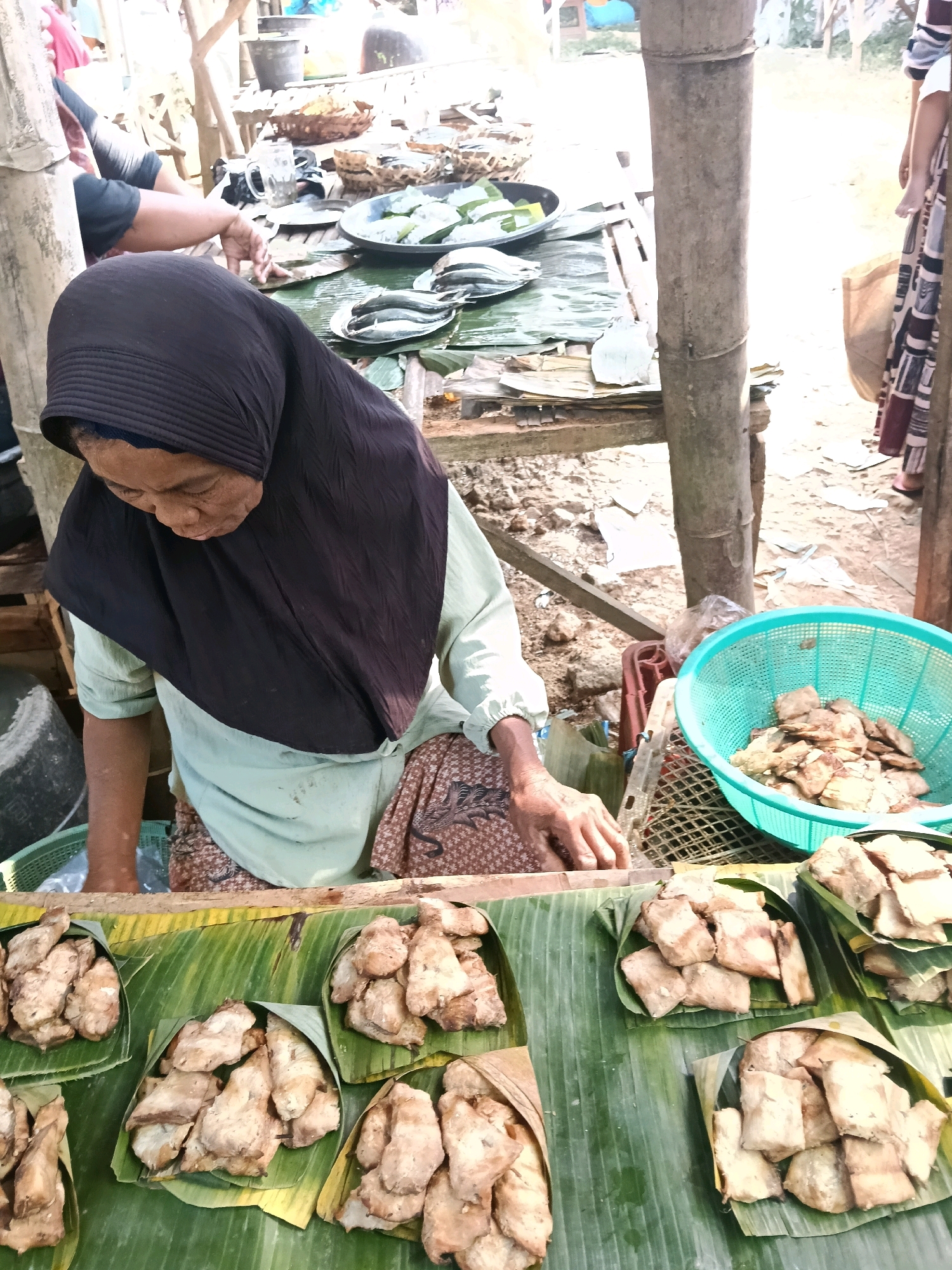 A vendor, a woman wearing a dark hijab and a light green shirt with a patterned skirt, stands behind a display of fried food, likely tempeh, arranged on banana leaves. The food is presented in portions wrapped in banana leaves and also piled in a turquoise plastic basket. The vendor's hands are visible, suggesting she might be arranging the food or interacting with a customer. In the background, other stalls with various items, including what appear to be fish on plates, and a basket of produce are visible. The scene appears to be an outdoor market with a rustic wooden structure in the foreground and a sandy ground. The lighting suggests it is daytime.