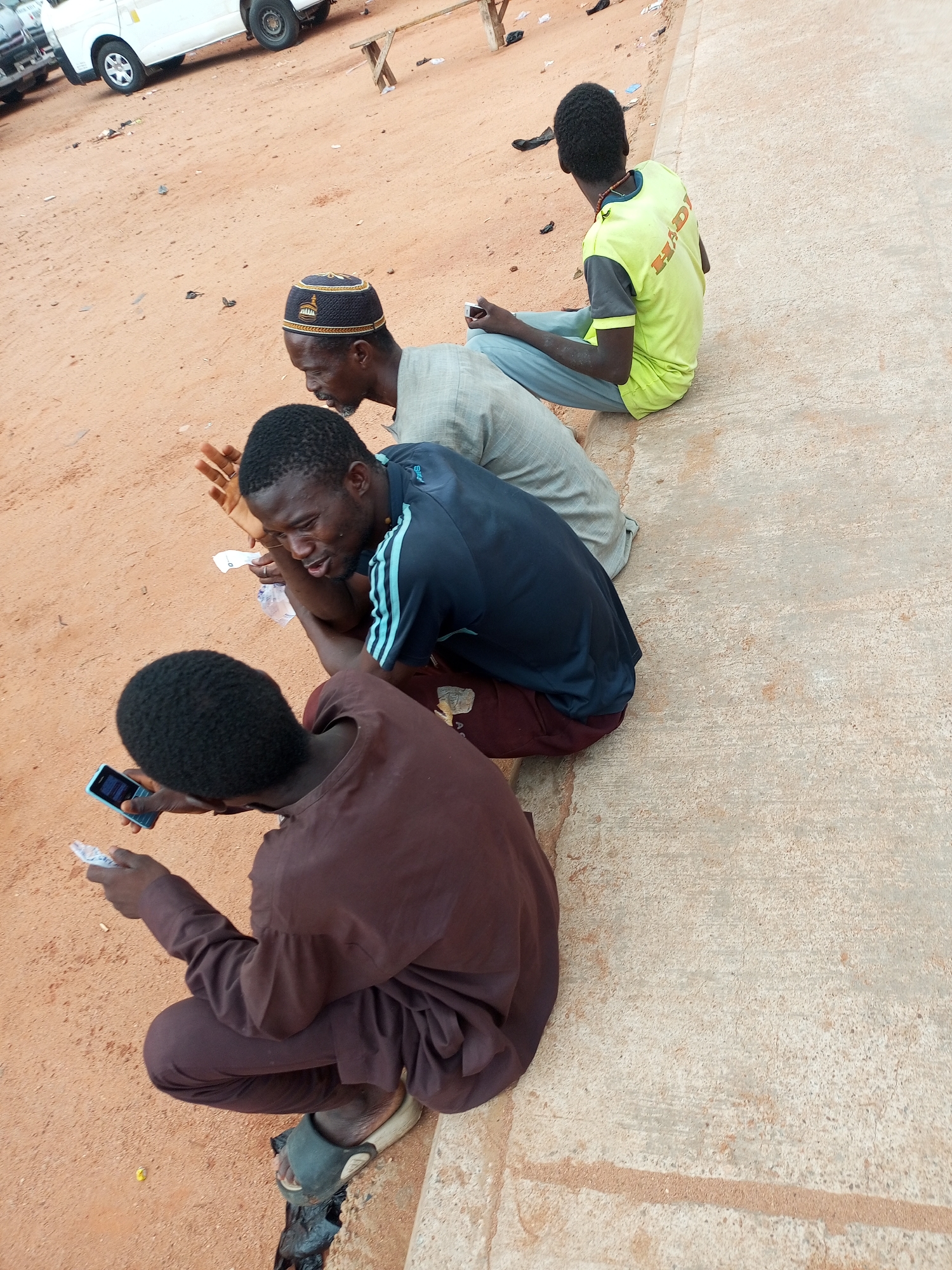 In the city of Gombe, Nigeria, four men are seen on a dusty, reddish-brown ground next to a concrete walkway. The man closest to the foreground, wearing a brown tunic and pants, is crouched and holding a light blue mobile phone in his left hand and a piece of white paper in his right. To his right, a man in a dark blue t-shirt with light blue stripes on the sleeves is leaning forward, looking down and gesturing with his right hand, a piece of white paper resting near his left arm. He is wearing burgundy pants. Behind him, an older man with a dark patterned cap is also looking down. He is wearing a light-colored tunic. The man furthest in the background, wearing a lime green and gray t-shirt and light blue pants, is sitting on the concrete walkway, looking away from the camera. A white van is partially visible in the upper left corner of the image, suggesting a location near a road or parking area. The ground is scattered with debris, including small pieces of white paper and dark plastic.