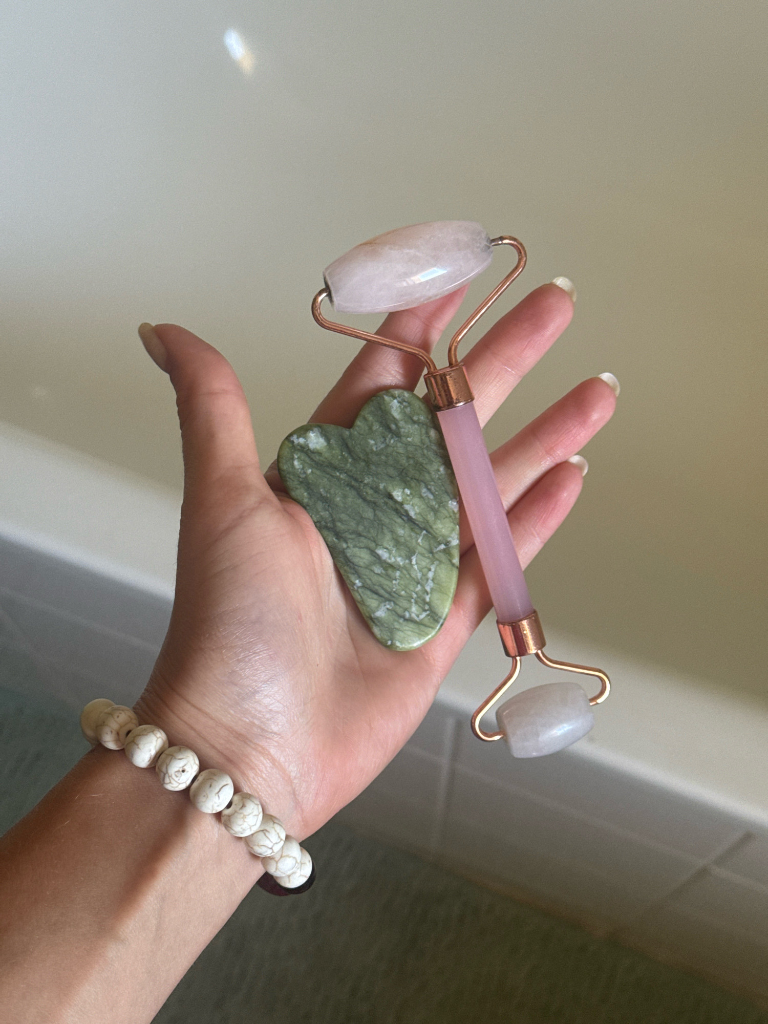 A person's hand, adorned with a white beaded bracelet, is held over a bathtub. The hand is holding a rose quartz facial roller and a green gua sha stone. The facial roller has a pink stem and rose gold accents, with a larger roller at the bottom and a smaller one at the top. The gua sha stone is heart-shaped and green with white veining. The background is a light-colored bathtub with a visible grout line. The lighting suggests this is an indoor scene. There is no information in the image to indicate the location in the city of Vienne, France.