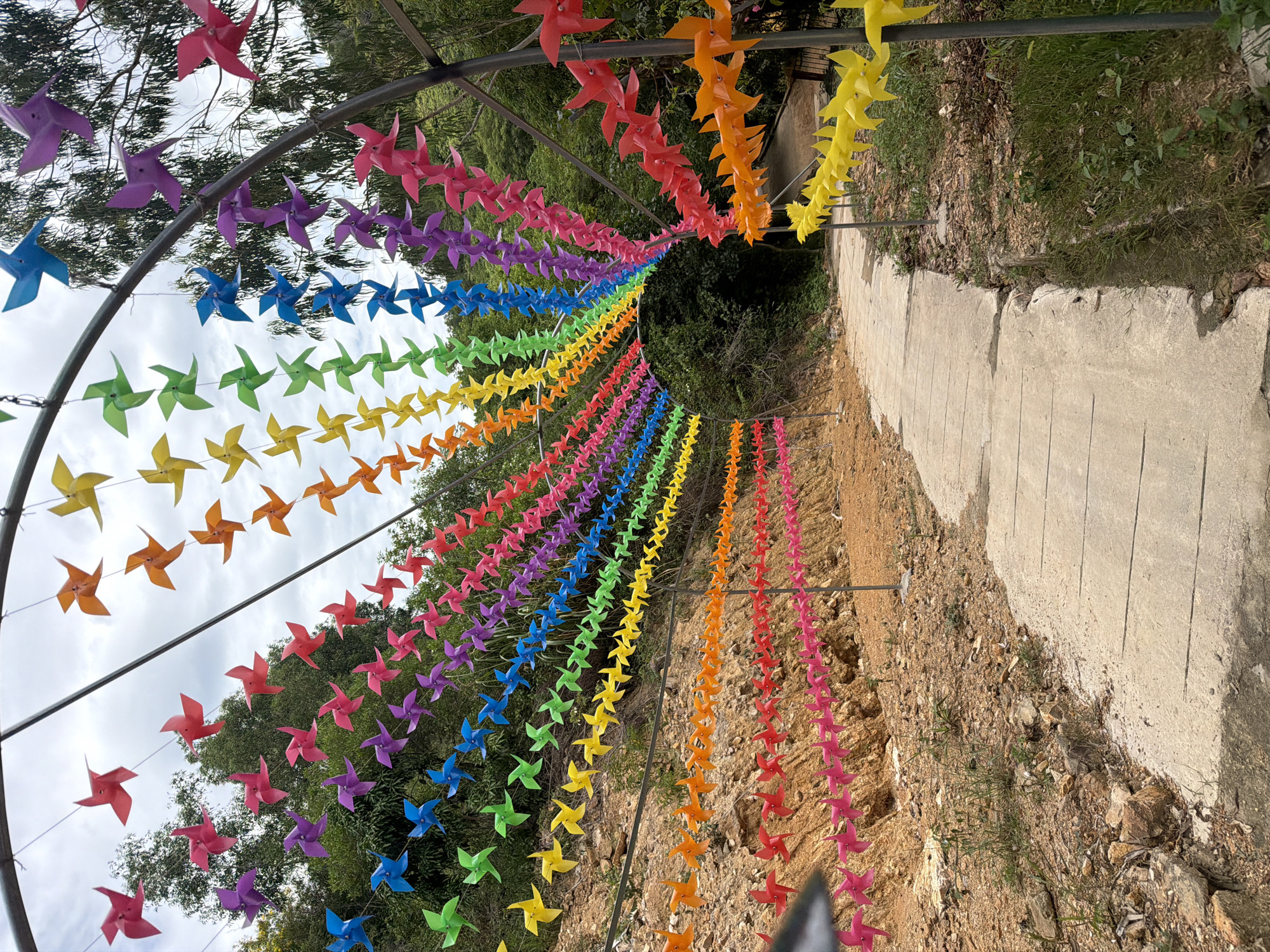 The image depicts a pathway lined with vibrant, multi-colored pinwheels forming an archway. These pinwheels are arranged in rows, cascading in a rainbow spectrum of colors: red, orange, yellow, green, blue, purple, and pink. They are attached to strings that are suspended from a metal arch structure, creating a decorative tunnel effect. The pathway itself is made of concrete slabs and curves to the right. The surrounding environment is a natural landscape, with a rocky, sloped terrain on the left side of the path, covered in sparse vegetation and dirt. On the right side of the path, there is denser greenery and trees. The sky is overcast with clouds. This scene is located at the entrance of the VinWonders Nha Trang amusement park on Hon Tre Island, Nha Trang, Vietnam. There are no people visible in the image.