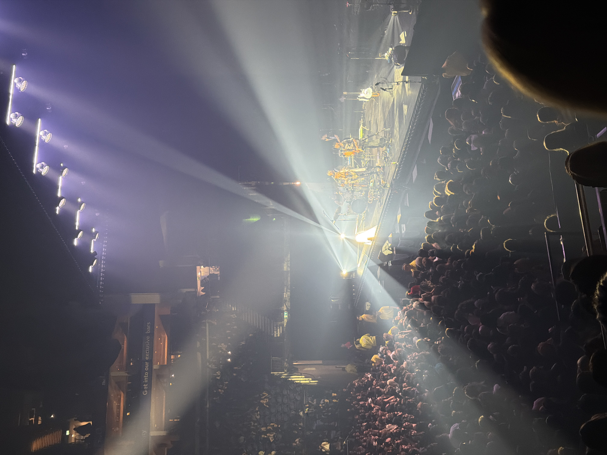 The image captures a concert scene from a high vantage point, possibly a balcony, within a large indoor venue. A dense crowd of people, their faces mostly obscured by shadow, is packed in front of a brightly lit stage where a band is performing. Light beams emanate from stage lighting, cutting through the air and illuminating sections of the crowd. To the left, a vertical sign reading "Get into our exclusive bars" is visible. The venue appears to be in London, United Kingdom. The image suggests a live music performance in a large entertainment arena.