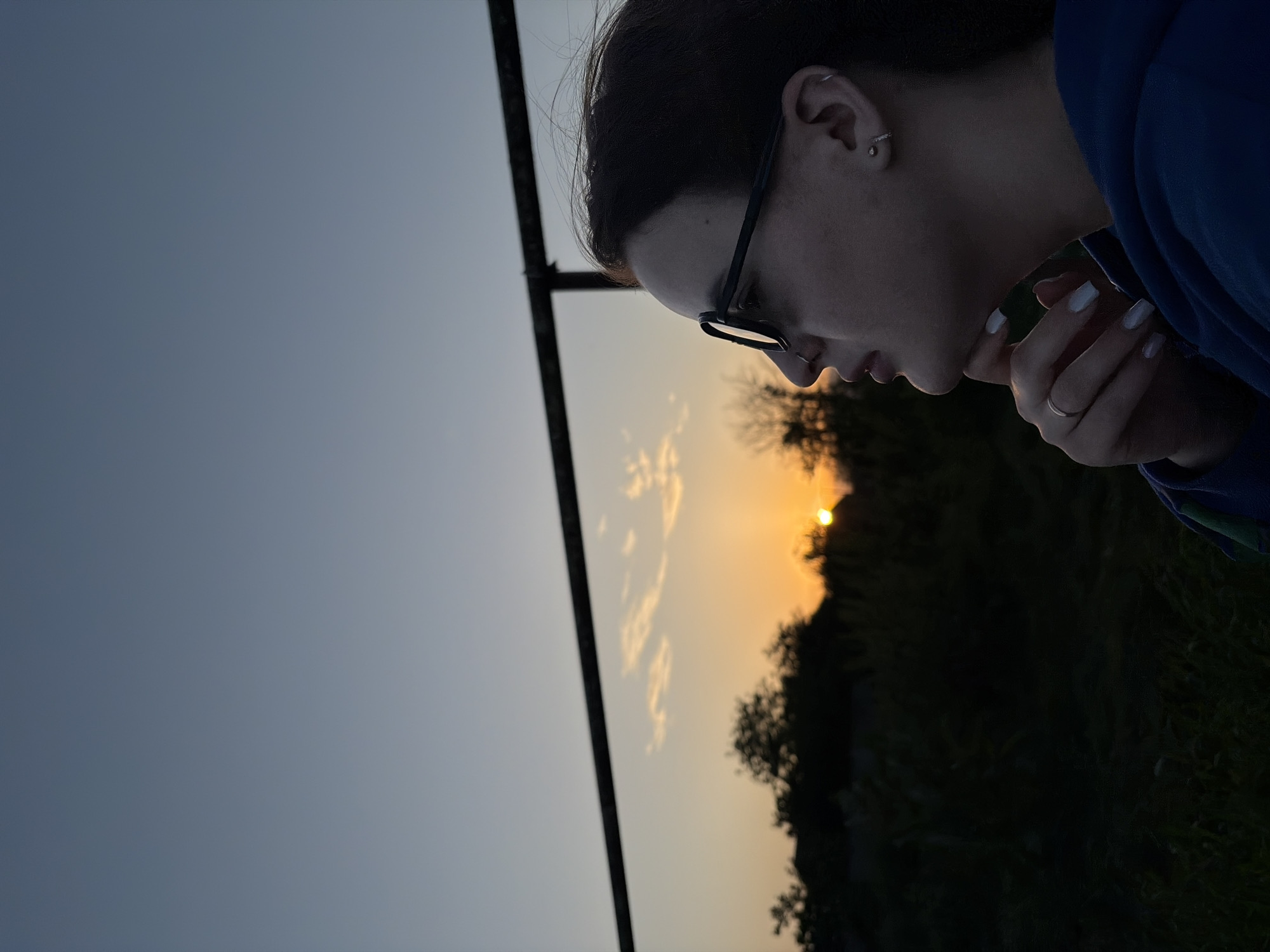A profile view captures a young woman with dark hair, wearing glasses, against a backdrop of a setting sun. She has fair skin, an ear piercing, and manicured white nails. She wears a blue garment. Her hand is near her face. Behind her, a fence is visible against the gradient sky, and silhouetted trees and vegetation. The sun is positioned near the horizon. The location is Novopokrovskaya, Russia.