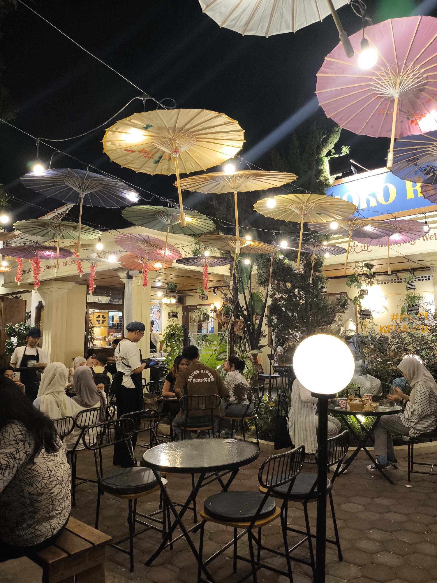 The image depicts an outdoor dining area at night in Surakarta, Indonesia, illuminated by hanging lamps and decorative paper umbrellas in various colors, including yellow, purple, and blue. The ground is paved with interlocking brick. Several individuals are seated at tables, some of whom appear to be dining. A waiter, wearing a white shirt and black apron, stands near a table. Another person, also in a white shirt and black apron, is serving food. Lush green plants are visible throughout the scene, contributing to a garden-like atmosphere. The establishment's signage, with blue lettering on a lighter background, is partially visible in the upper right quadrant. The overall ambiance suggests a lively evening gathering at a restaurant.