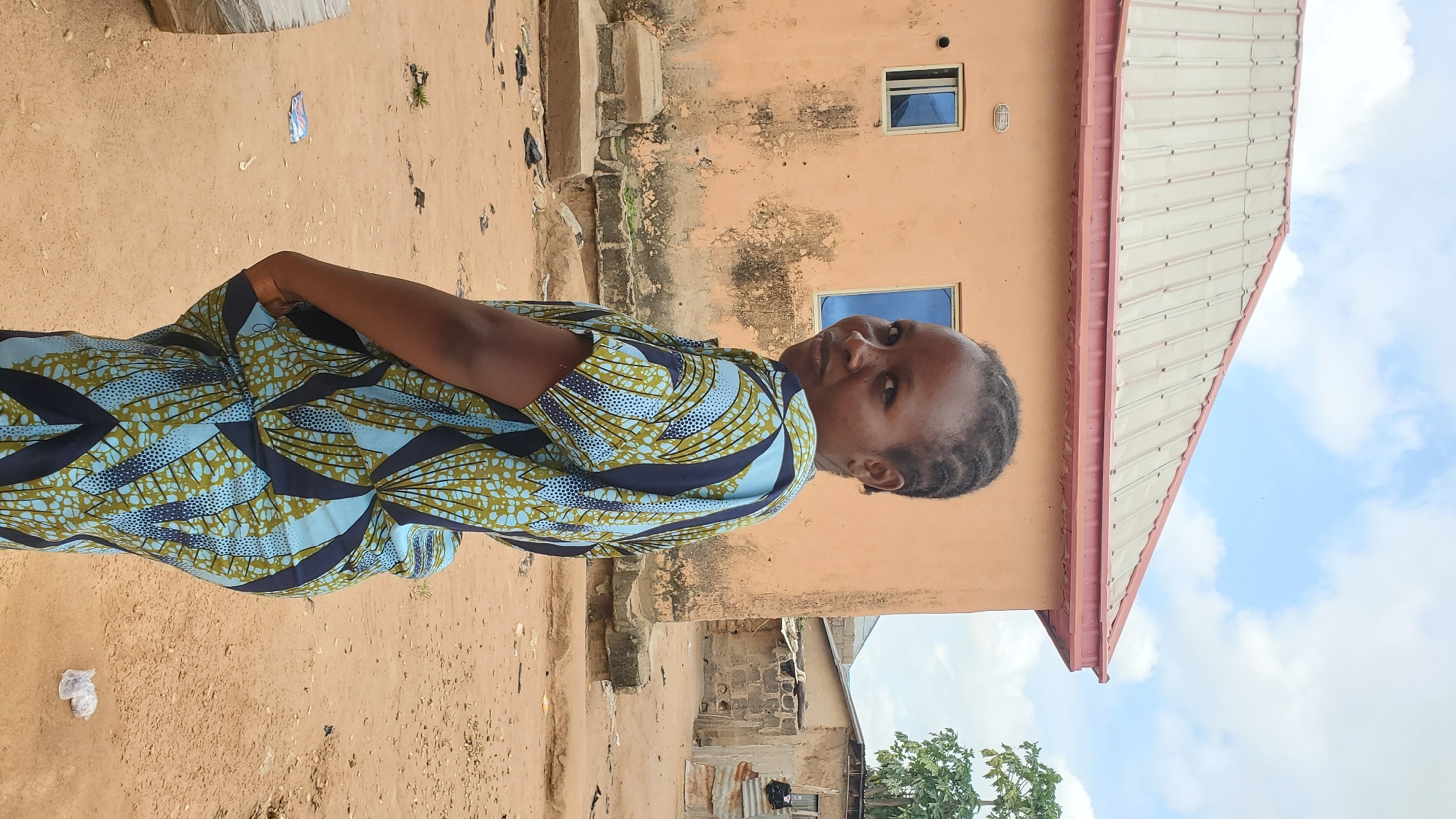 A medium shot captures a woman with dark skin, dark braided hair, and dark eyes standing outdoors in a village setting. She is positioned at a slight angle to the camera, looking back over her left shoulder with a neutral expression. She is wearing a vibrantly patterned dress in shades of blue and green with a dark navy blue base and an all-over design of abstract shapes and circles. The dress has short sleeves and appears to be made of a lightweight fabric.
Behind the woman, an orange building with a corrugated metal roof is visible. The building has two rectangular windows with blue panes. The ground in the foreground and background is a sandy, reddish-brown dirt, with scattered debris including plastic wrappers and what appear to be a pair of black flip-flops. In the distance, other buildings constructed with various materials, including corrugated metal and what looks like mud brick, are visible. A tree with green foliage stands to the right of the frame, partially obscuring some of the background structures. The sky above is a pale blue with scattered white clouds.
The image appears to have been taken in the city of Jalingo, Nigeria, a location known for its rural and semi-rural communities where such housing and environmental conditions are common.