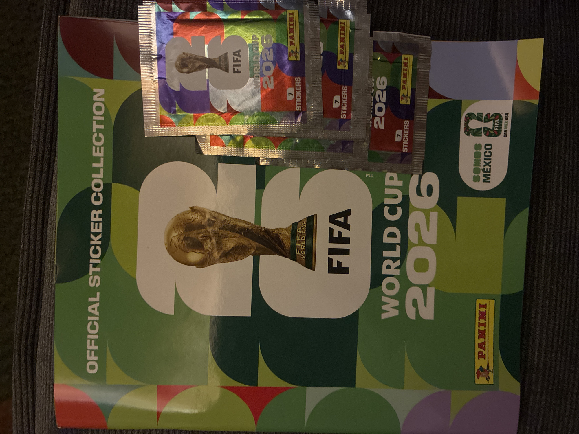 An overhead shot displays a Panini FIFA World Cup 2026 Official Sticker Collection album and several sealed sticker packs, presumably in Huixquilucan de Degollado, Mexico. The album, serving as the main subject, features a vibrant green cover adorned with geometric shapes in varying shades of green, purple, red, and light blue. A prominent graphic of the FIFA World Cup trophy dominates the center, with "FIFA WORLD CUP 2026" text beside it. The title "OFFICIAL STICKER COLLECTION" runs vertically along the left edge. Towards the bottom right, a smaller logo reads "SOMOS MÉXICO" and "FIFA CAN MEX USA," indicating a focus or release related to the host nations. The Panini logo is visible on the album and sticker packs.
Stacked atop the album are multiple metallic, colorful sticker packs, each repeating "FIFA WORLD CUP 2026" and clearly stating "7 STICKERS." The items are arranged on a dark, textured surface, possibly a blanket or carpet, under what appears to be indoor, somewhat dim lighting. No human subjects or explicit activity are captured, but the scene implies the collector's pastime of gathering World Cup stickers.