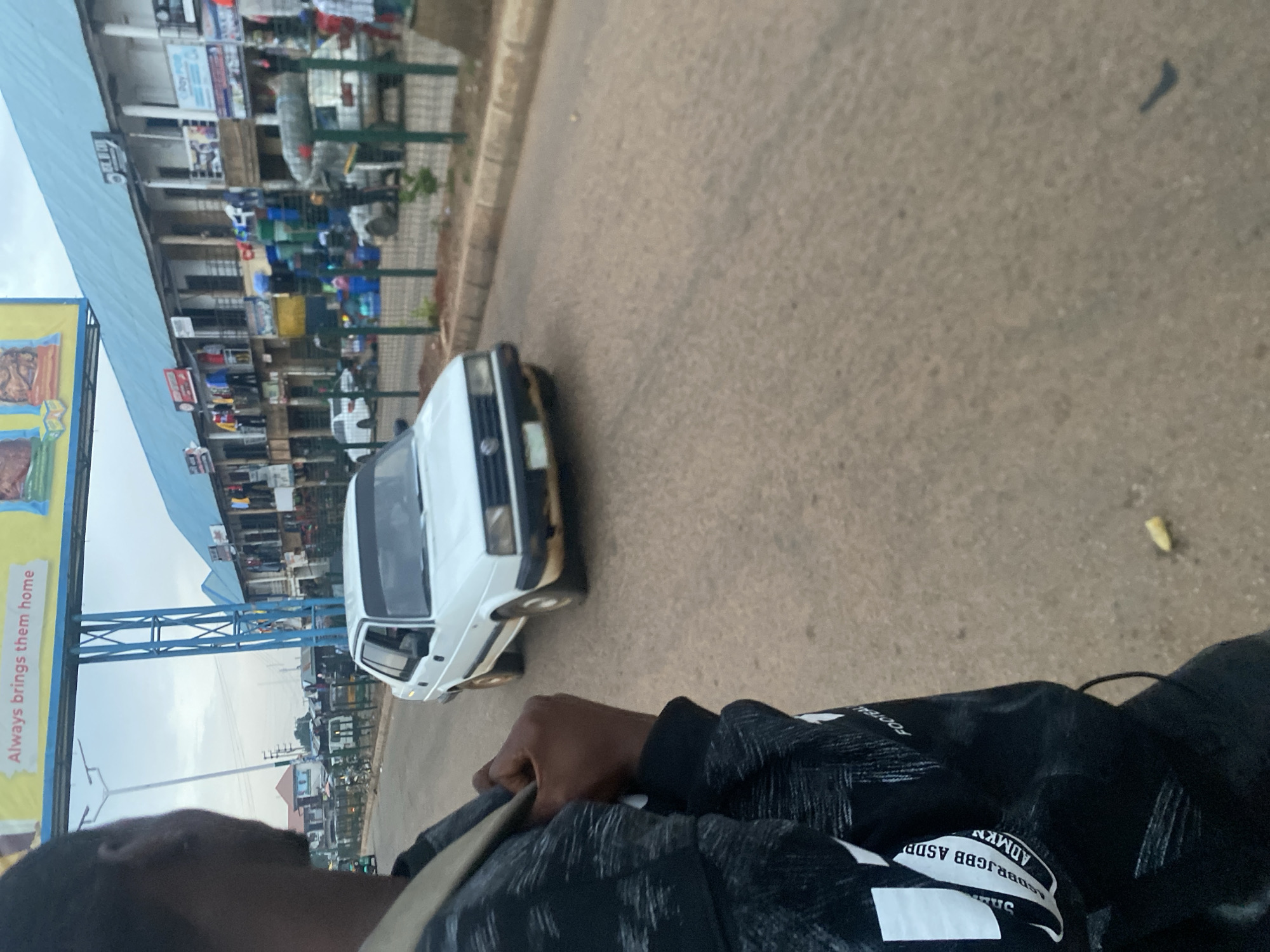 A medium-shot, eye-level, outdoor, daylight scene captures a street in Bukuru, Nigeria. In the foreground, a person's left shoulder and back are visible, draped with a black jacket featuring a white pattern and text. The person is looking forward, with their left hand grasping the jacket's strap.
Dominating the mid-ground is a white Volkswagen car, its front facing the viewer's left. The car is on a paved road. Beyond the road, a row of two-story buildings forms a marketplace. These buildings have light blue roofs and balconies. Numerous stalls and shops are visible on the ground floor, displaying various goods, including clothing, plastic containers, and what appears to be food items. Green fencing separates the road from the marketplace.
In the background, a large blue metal structure supports a billboard with yellow and white text. The sky is overcast. The overall atmosphere suggests a typical urban environment in Nigeria.