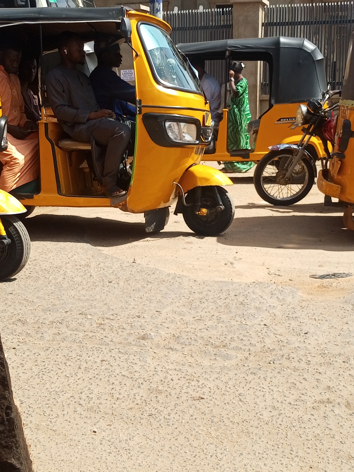 The image depicts a row of yellow auto rickshaws, locally known as kekes, on a paved street in Maiburiji, Nigeria. In the foreground, one keke is occupied by four passengers and a driver. The driver wears a dark shirt, and one passenger wears an earpiece. Another keke is visible in the mid-ground, with a passenger in green patterned clothing. Metal gates are visible in the background. The foreground shows details of the paved road.