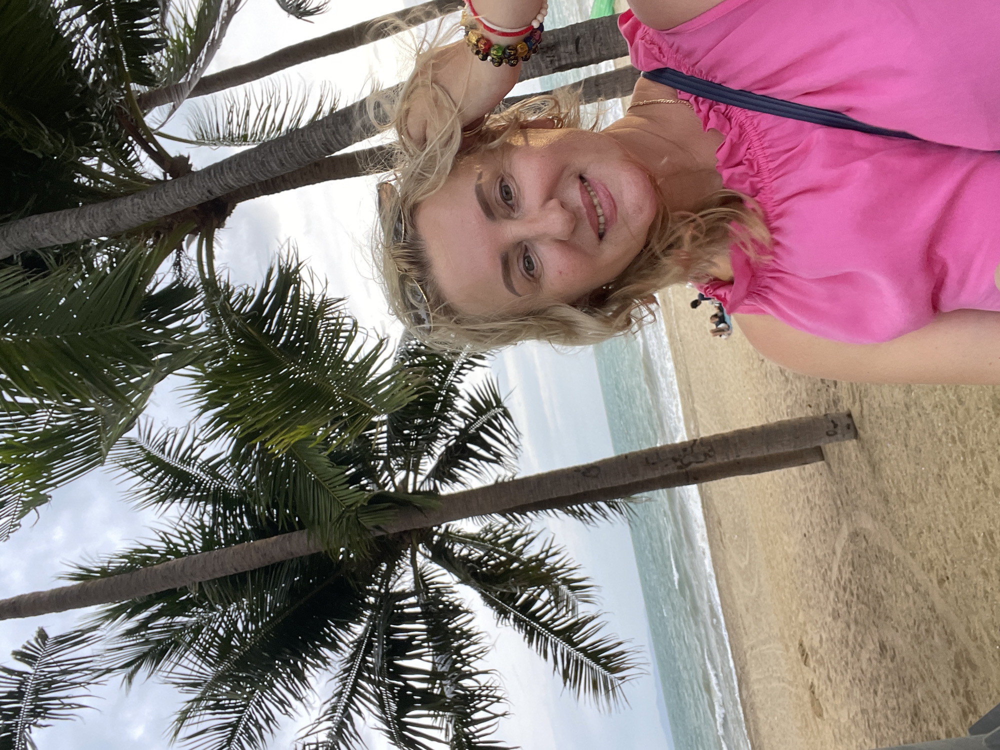 A woman takes a selfie on a beach in Pattaya City, Thailand. She is wearing a pink top and has blonde hair. Palm trees are visible in the background, as well as the ocean and the sand. There are a few other people on the beach in the distance. The sky is cloudy.