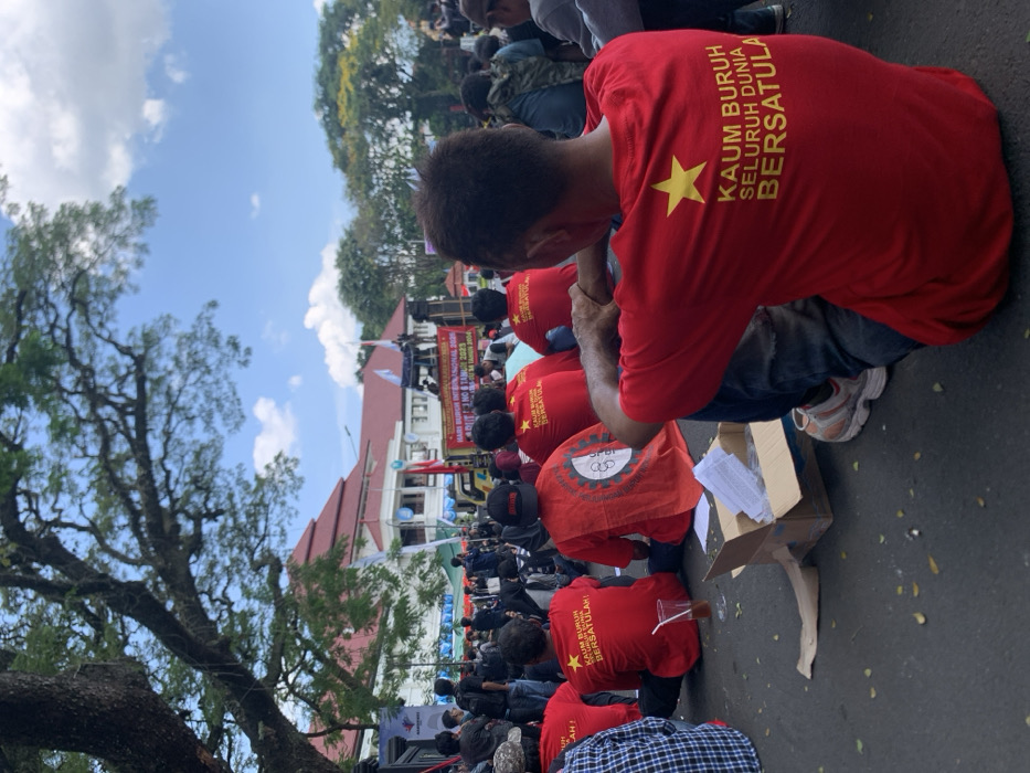 The image shows a group of people sitting on the ground in front of a building in Malang, Indonesia, located at coordinates -7.977 latitude, 112.634 longitude. They are all wearing red shirts with yellow stars and slogans in Indonesian, including "KAUM BURUH SELURUH DUNIA BERSATULA," which translates to "Laborers of the Whole World Unite." Some are wearing hats, and some are holding signs. There is a tree in the background. There is a large banner in the background that says, "HARIBO BURUH INTERNASIONAL SE-ASIA - 1 NO - 8 TAMAN 2023." The individuals are gathered around a cardboard box and are seated on a paved area. The scene is likely a labor protest or rally. It appears to be a peaceful gathering.