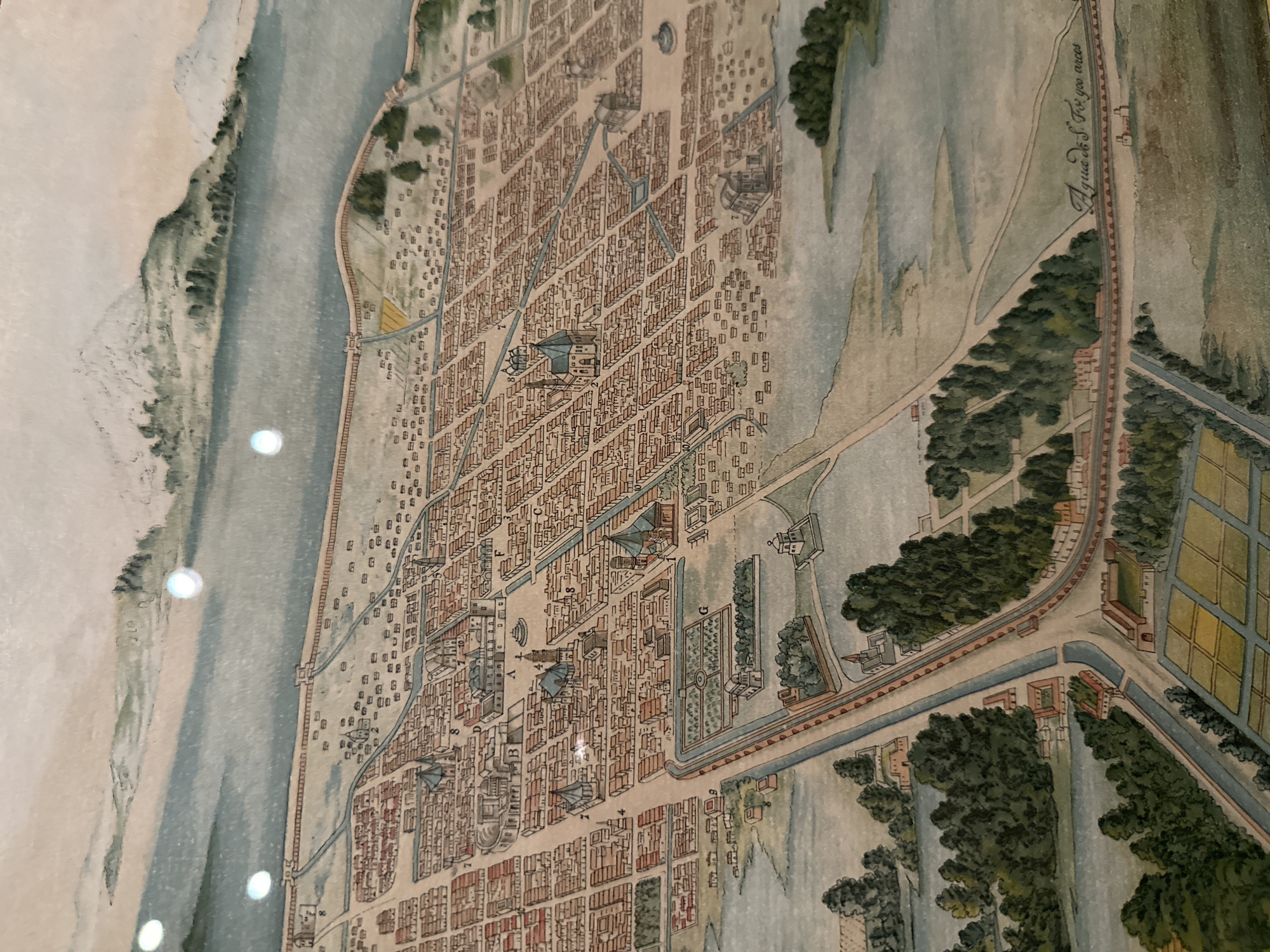The image shows an antique map of Mexico City. It depicts a densely packed urban area characterized by closely arranged buildings, waterways, and roads. The river runs alongside the city's border, with a clear difference between the developed area and the surrounding landscapes. The map indicates areas such as parks or gardens, with distinct details. In the upper right corner of the image, the inscription "Agua de S. Fo yoo arces" can be observed.