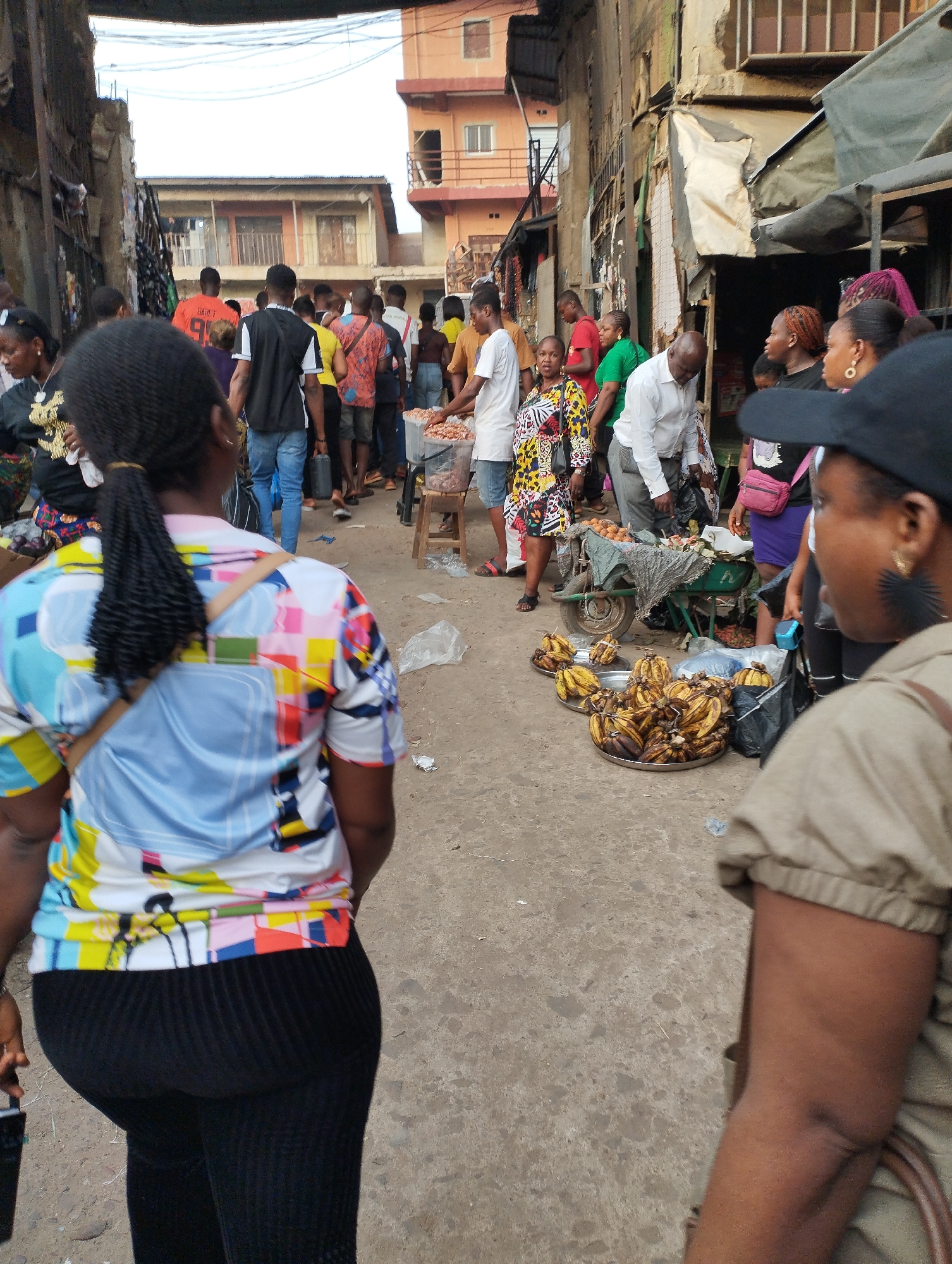 The image captures a bustling street scene in Fegge, Nigeria. A large group of people is milling about on a narrow, concrete street lined with buildings. Many of the buildings appear aged, with visible weathering and some makeshift awning structures. The scene is filled with activity as people are shopping and interacting with vendors.
In the foreground, there are bananas spread out on platters on the ground, presumably for sale. A woman wearing a colorful patterned top and black pants stands with her back to the camera. Another woman in a khaki shirt and baseball cap stands on the right. In the middle ground, several vendors are selling items. Further down the street, a person standing on a stool appears to be selling groundnuts, judging by the containers nearby. In the background, more buildings can be seen, giving depth to the urban setting. Overall, the image portrays a vibrant and crowded commercial area.