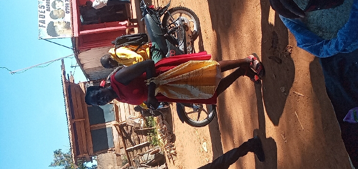 The image shows a woman in a red dress standing next to a motorcycle in Lwandeti, Kenya, located at latitude 0.563 and longitude 34.806. The woman is holding a yellow and white bag. Another man, wearing a yellow shirt and a black backpack, is sitting on the motorcycle. The motorcycle is parked in front of a barber shop. There are wooden buildings on the left side of the image. The ground is dirt. The woman's shadow is cast on the ground. There is a partially visible person in the foreground wearing a blue jacket. The sky is blue and cloudless.