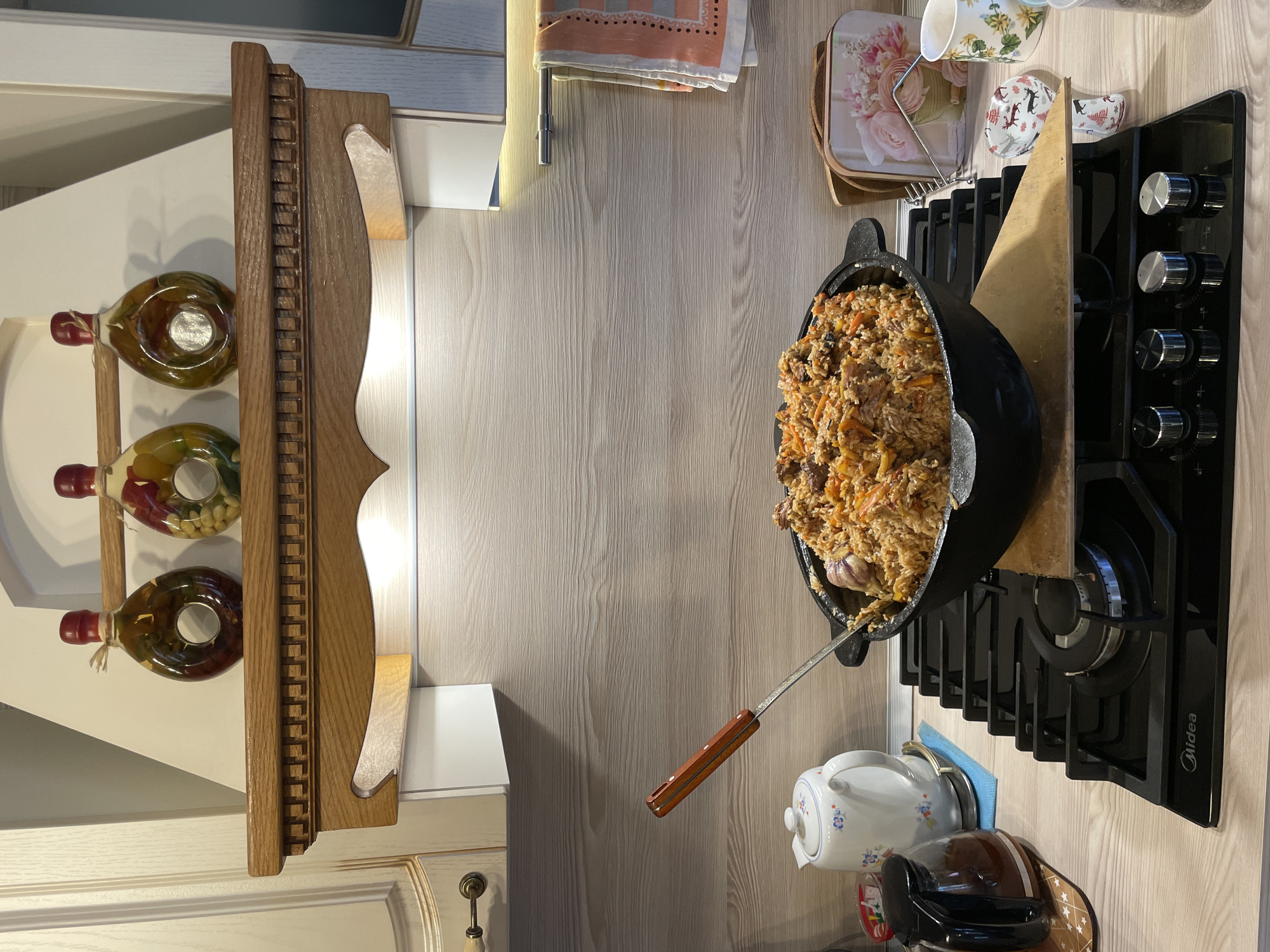 The image shows a kitchen counter with a black pot of pilaf on a gas stove. The pot is large and round, and it is filled with rice, meat, and vegetables. A metal spoon with a wooden handle is sticking out of the pot. To the left of the stove, there is a wooden shelf with three bottles of oil and spices. Above the stove, there is a countertop with some dishes and utensils. The location of the scene is likely in Ленинградская, Russia.