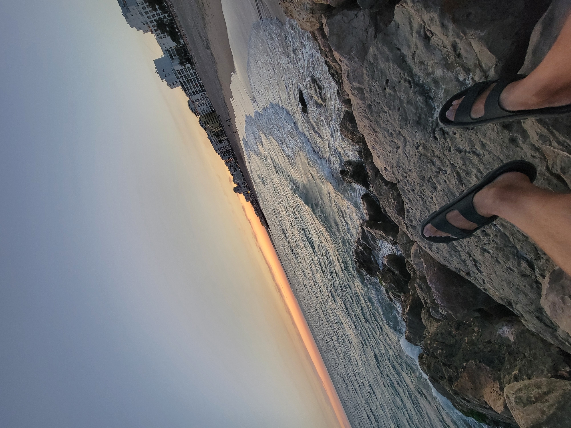 The image depicts a low-angle perspective of a rocky shoreline overlooking the ocean. In the foreground, a person's legs and feet, wearing dark sandals, are visible, positioned on large, gray rocks. The ocean is a deep blue with white-capped waves crashing against the rocks. The shoreline extends to the right, revealing a sandy beach lined with white buildings and palm trees. The sky is a pale blue, transitioning to hues of orange and yellow near the horizon, indicating either sunrise or sunset. The overall scene is one of a coastal environment in Quarteira, Portugal.